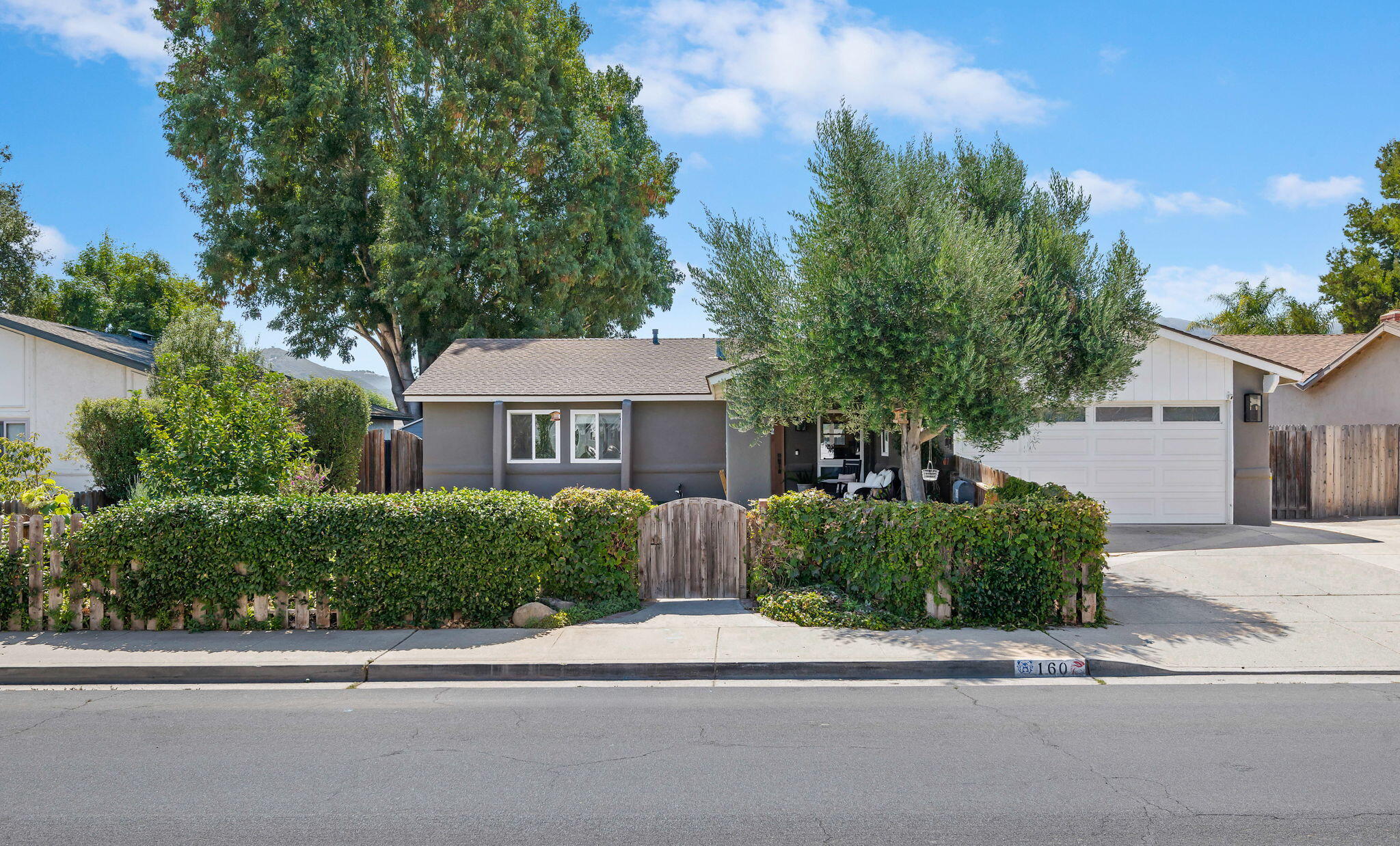 Single-story gray house with a white garage, mature trees, and a hedge-lined front yard.