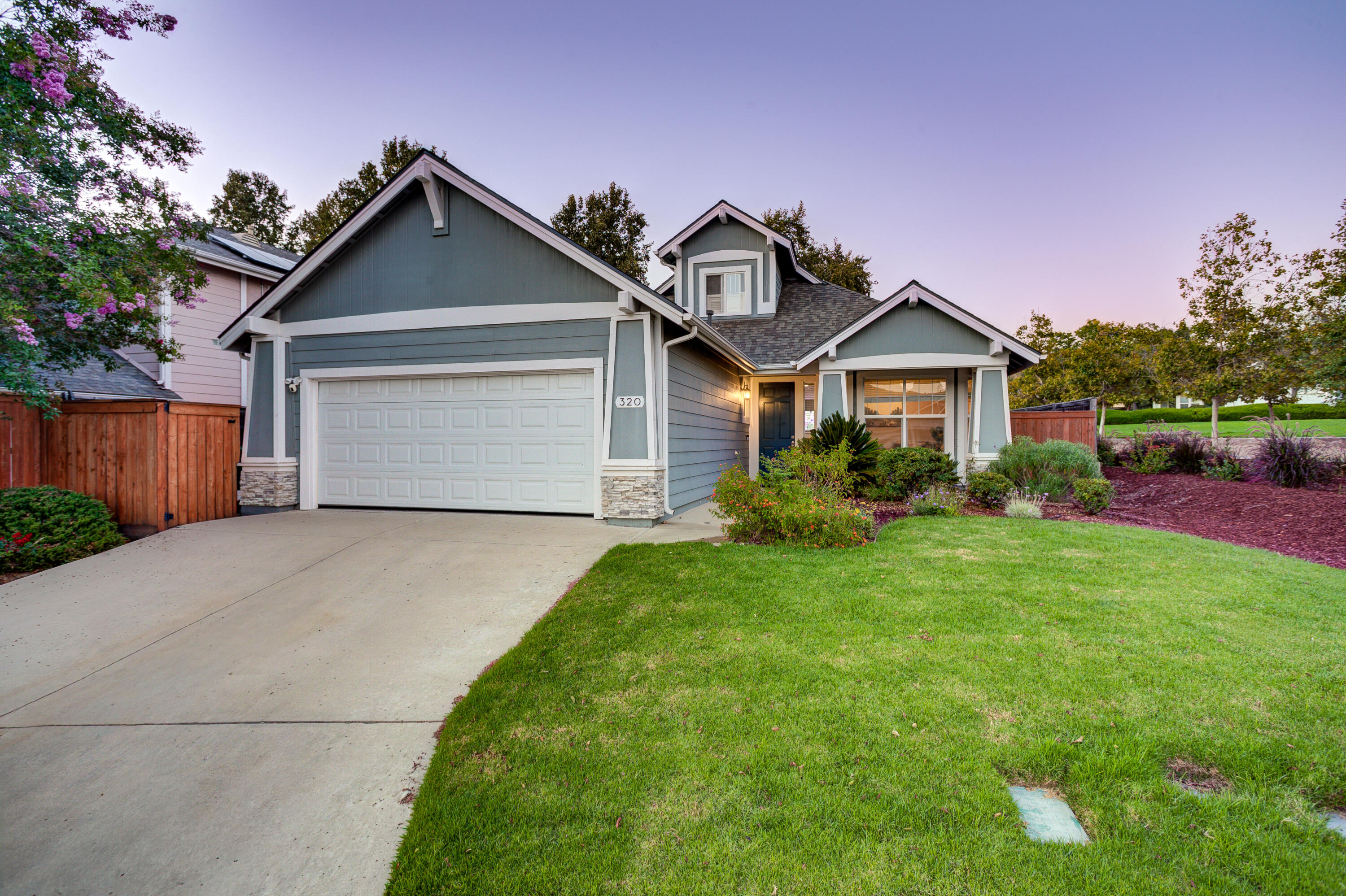 Gray-blue gabled home with a white double garage, broad driveway, and landscaped front lawn at dusk.