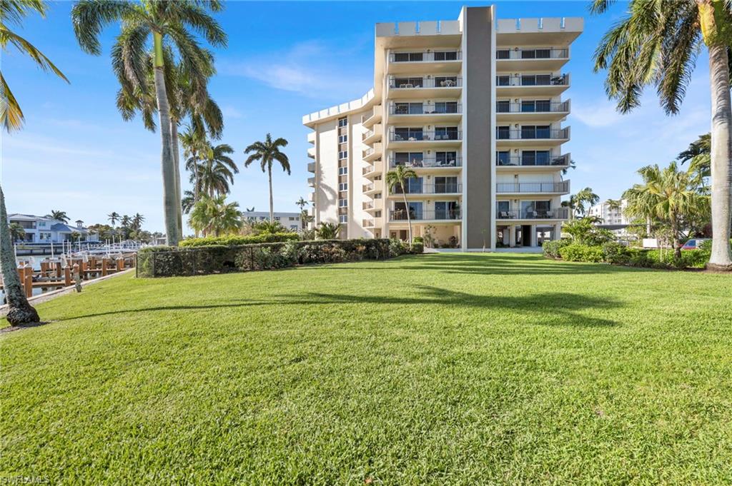 Condominium building with stacked balconies behind a broad lawn, framed by palms beside a waterfront dock.