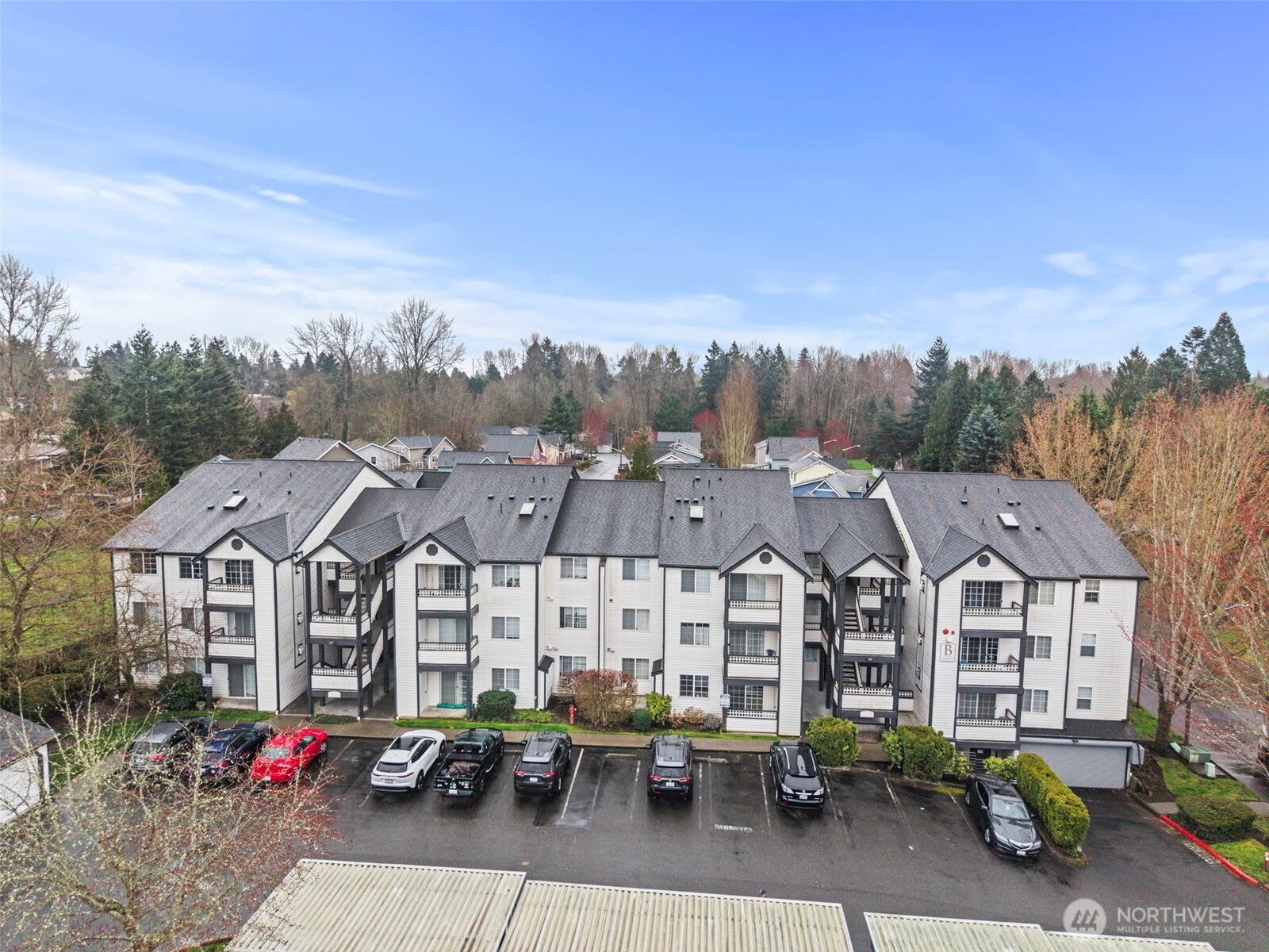 Aerial view of a white multi-unit residential building with balconies, parking, and wooded surroundings.