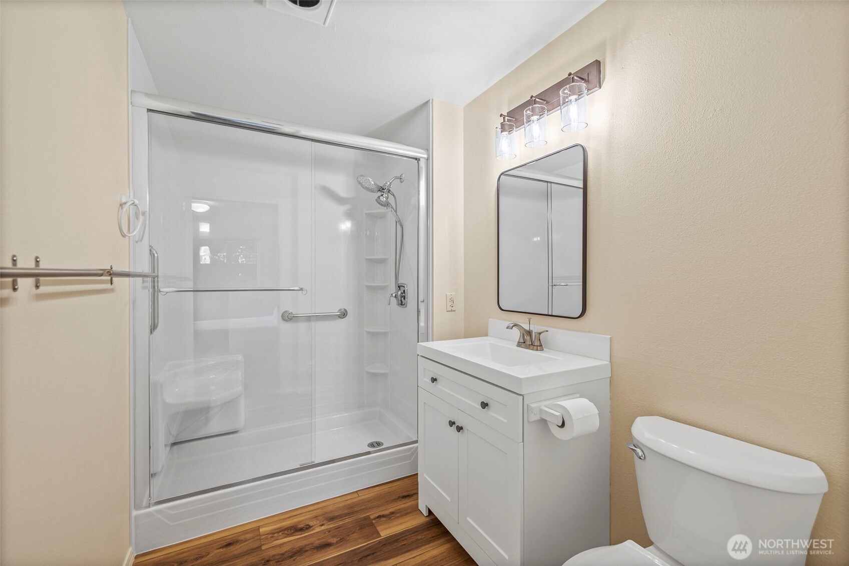 Bathroom with a glass-enclosed shower, built-in seat and grab bars, white vanity, and wood-look flooring.