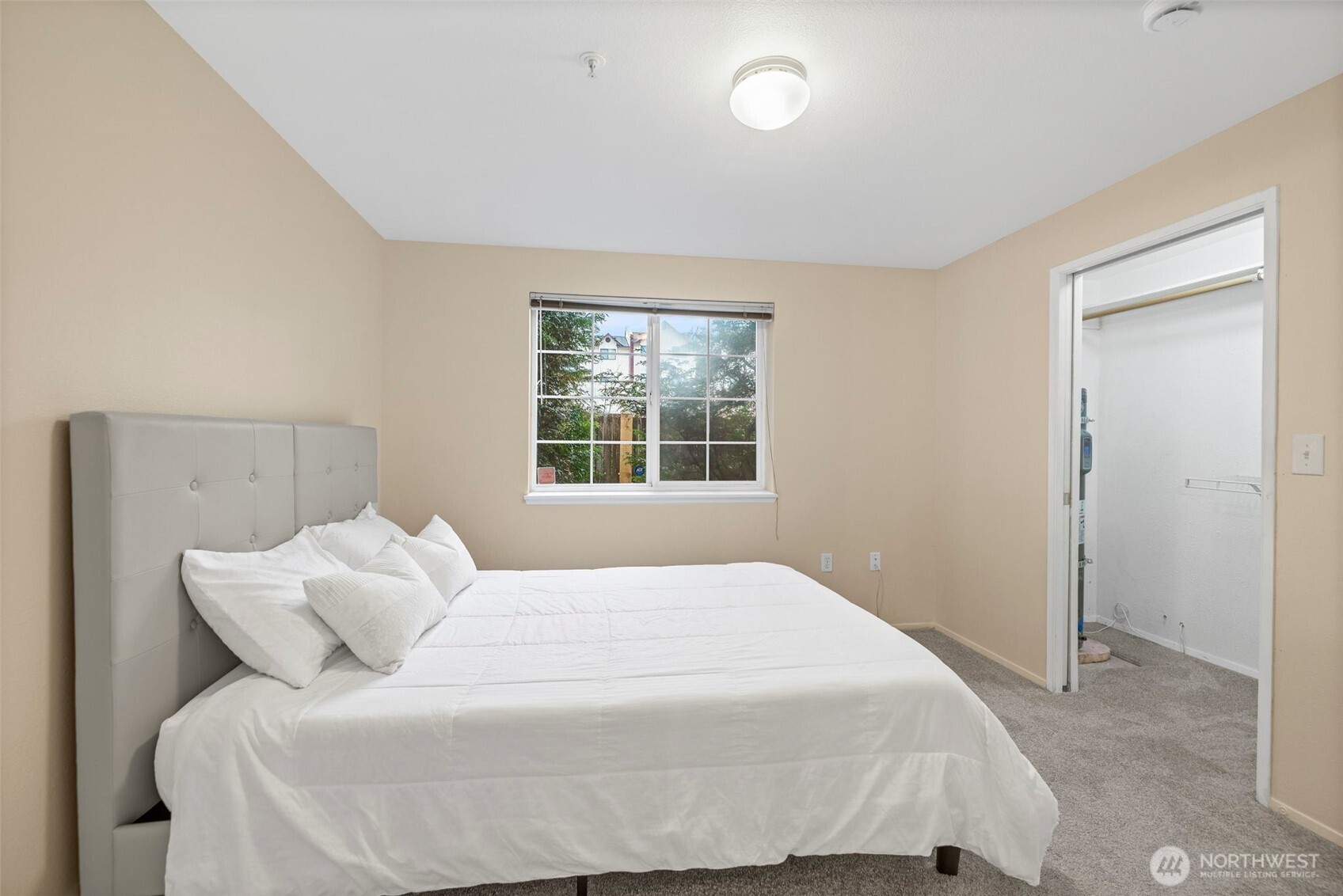 Staged bedroom with a white bed, gray upholstered headboard, and window overlooking greenery.