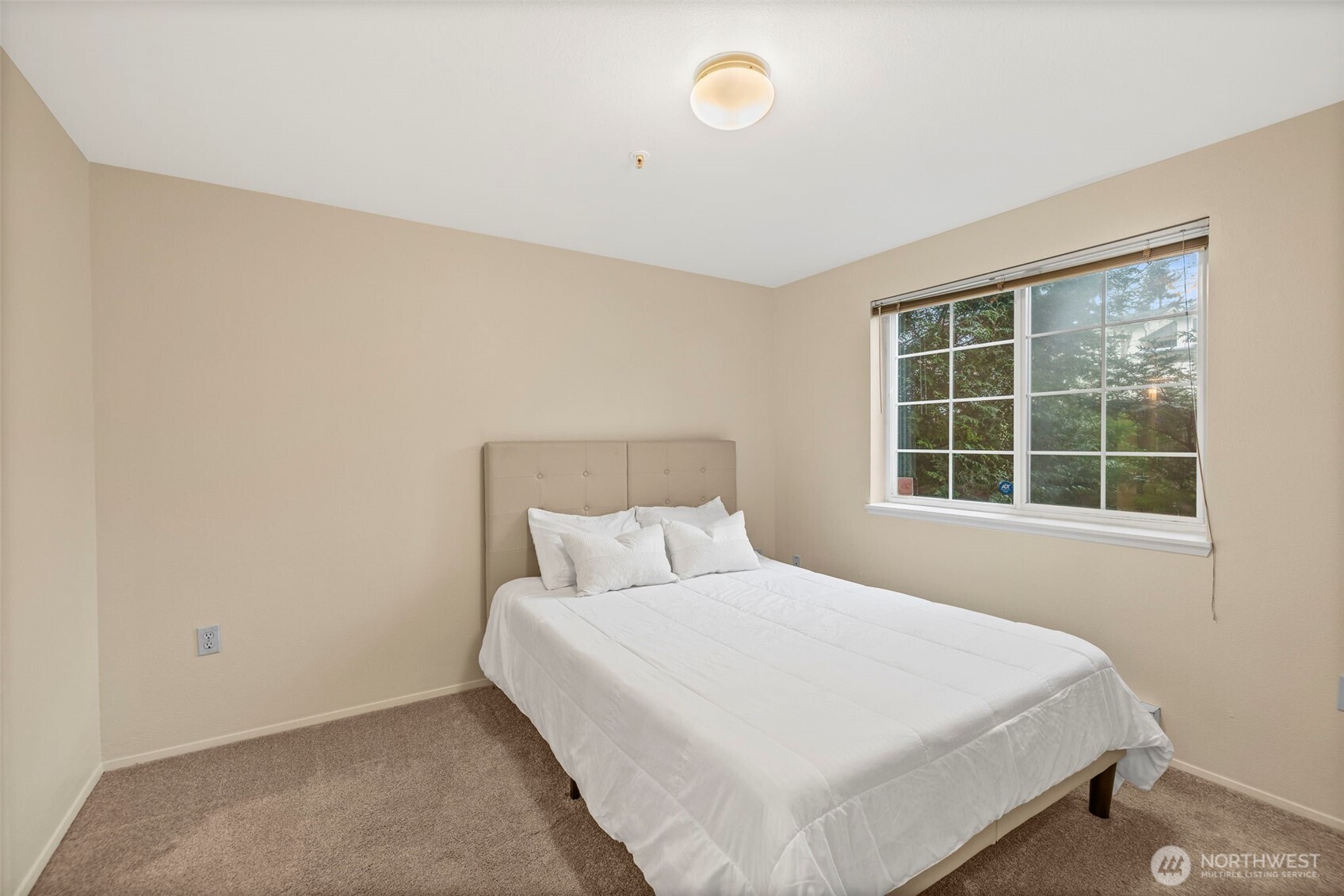 Bedroom with a white bed, beige tufted headboard, brown carpet, and a large window overlooking trees.