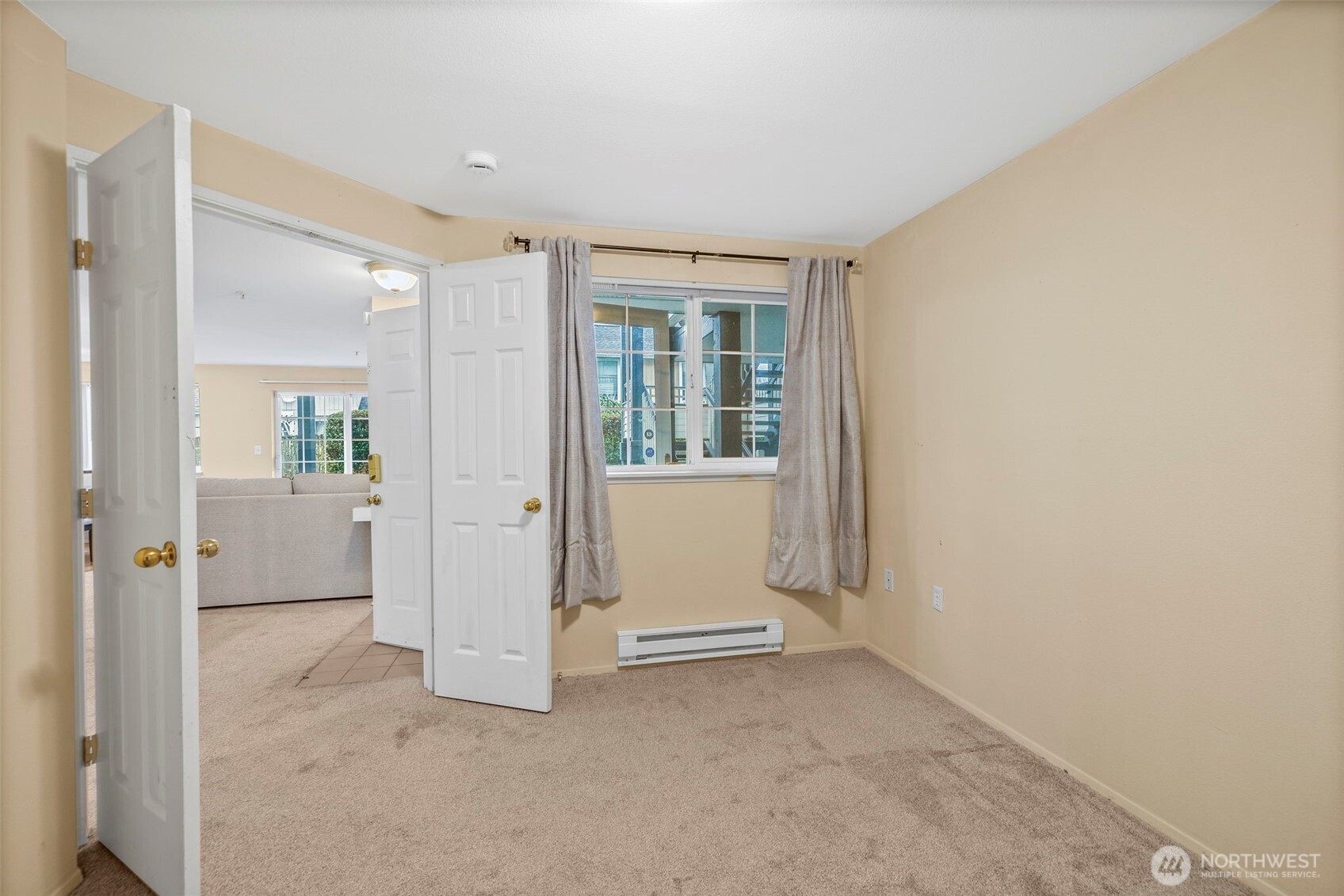 Empty carpeted room with beige walls, gray curtains, and open doors to a living area.