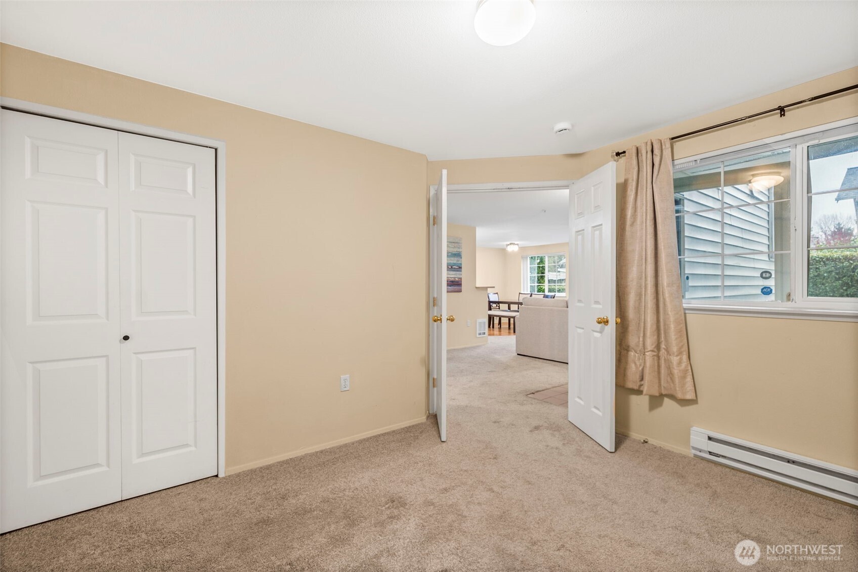 Empty carpeted bedroom with sliding closets, a curtained window, and an open doorway to the living area.