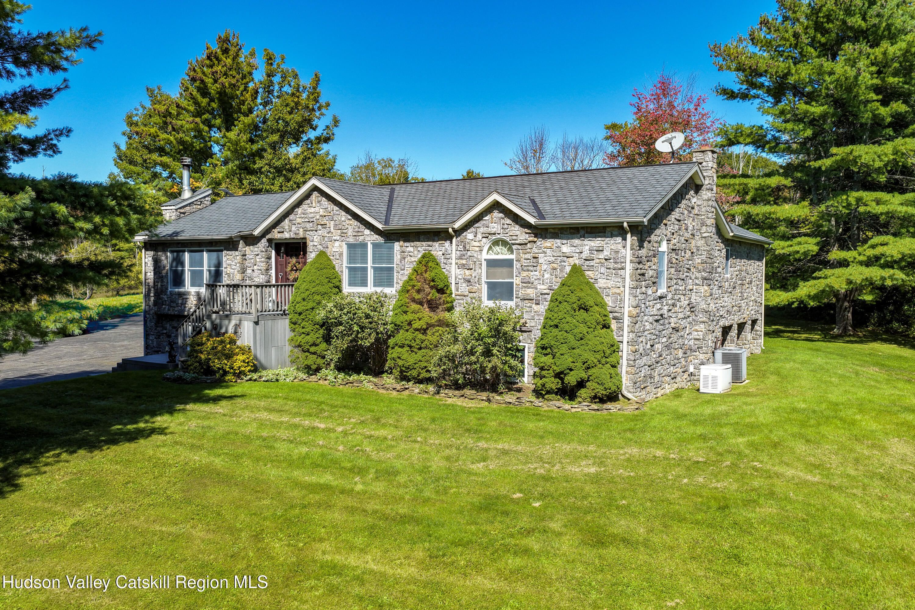 Stone house with a gray shingle roof, front deck, and landscaped lawn surrounded by trees.