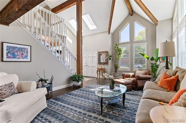 Vaulted living room with exposed beams, a white staircase, tall windows, and seating around a glass coffee table.
