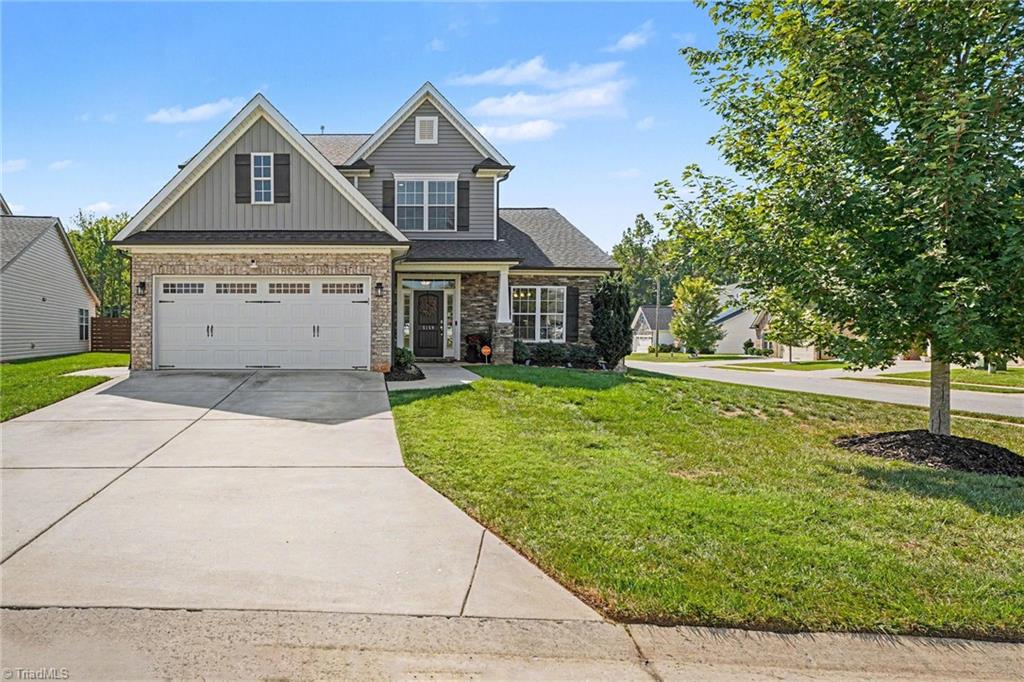Two-story gray house with stone accents, double garage, wide driveway, and a mature tree in the front yard.