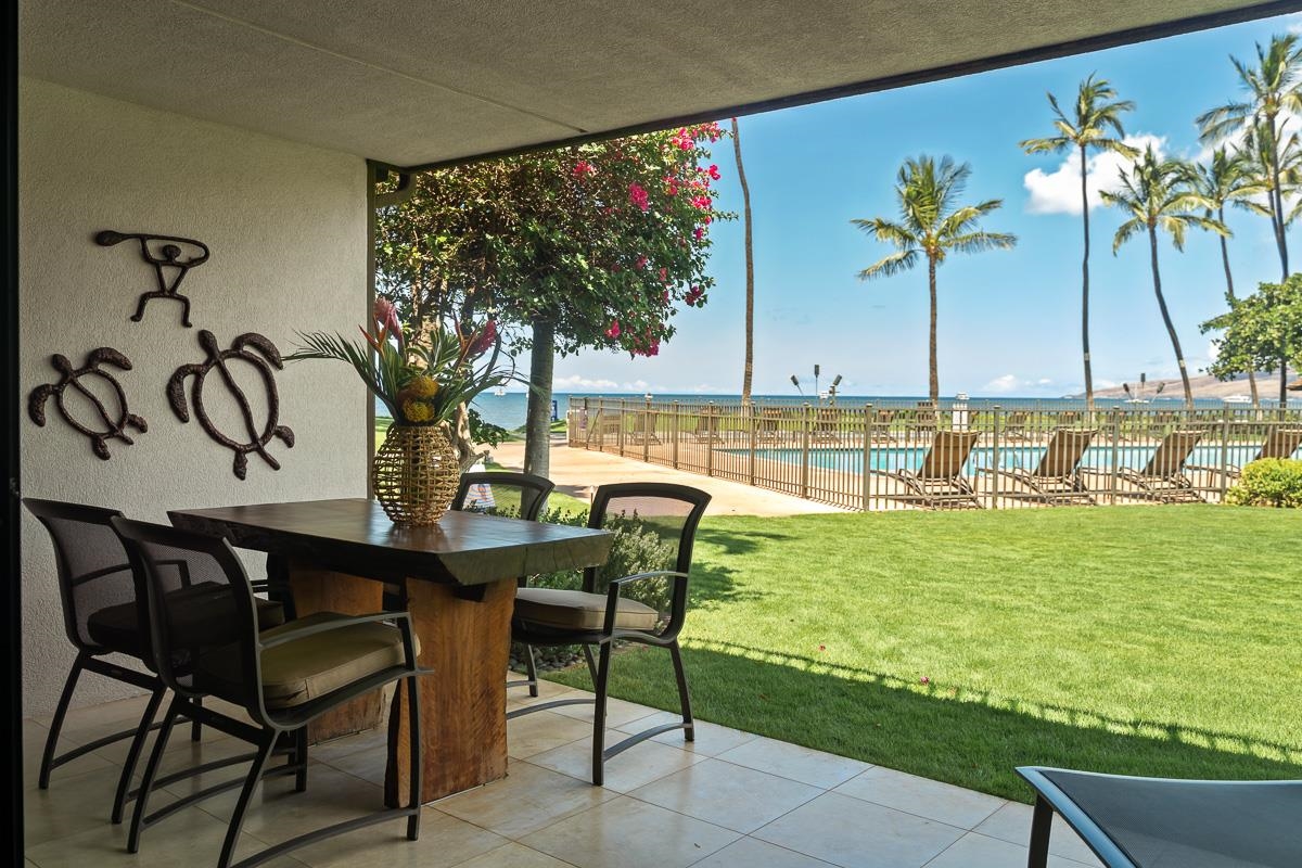 Covered patio dining area overlooking a lawn, pool, palm trees, and ocean.