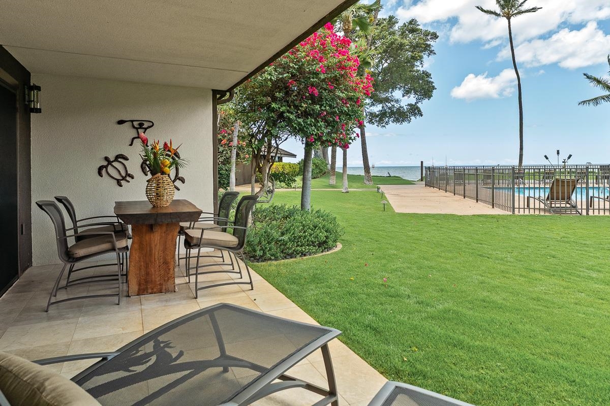Furnished patio overlooking a lawn, pool and ocean, with a flowering tree nearby.
