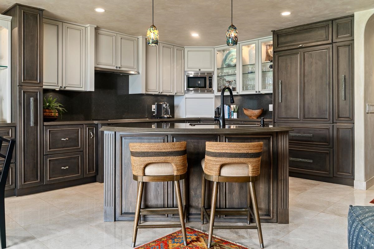 Kitchen with a dark wood island, two woven barstools, pale cabinetry, and glass-front cupboards.