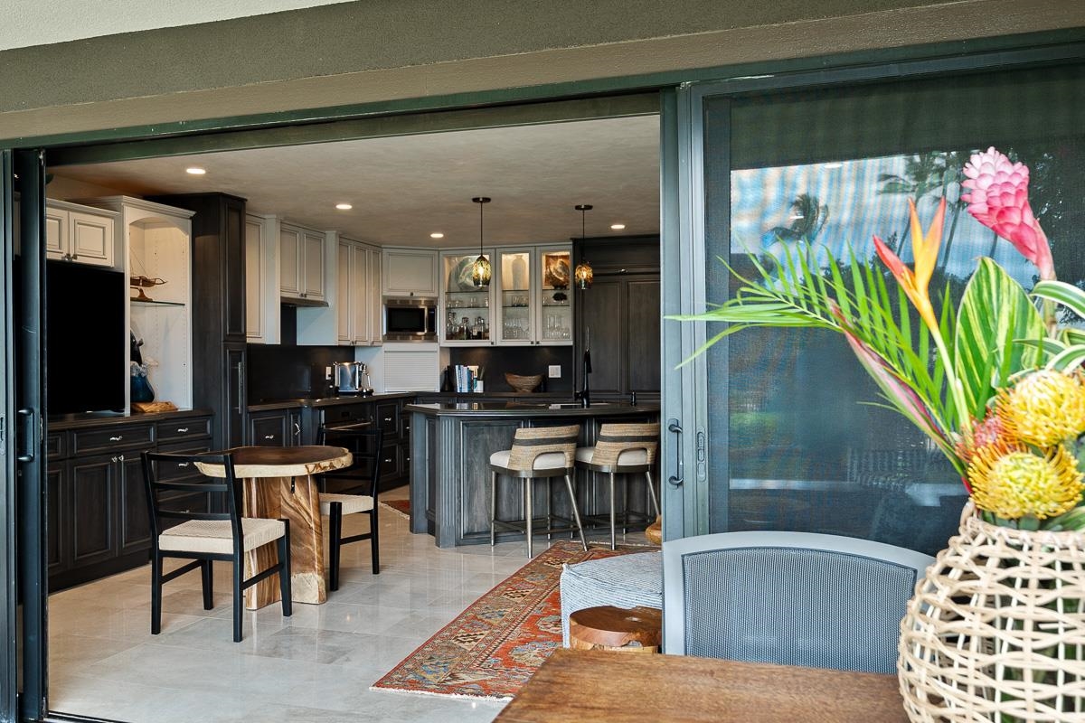 Furnished kitchen with a dark wood island and bar stools, viewed past tropical flowers by a sliding door.
