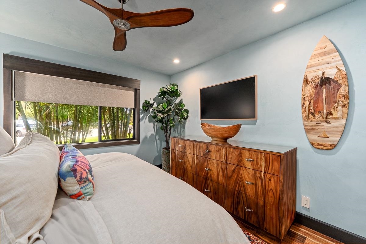 Bedroom with a white bed, wood dresser, ceiling fan, and palm trees visible through the window.
