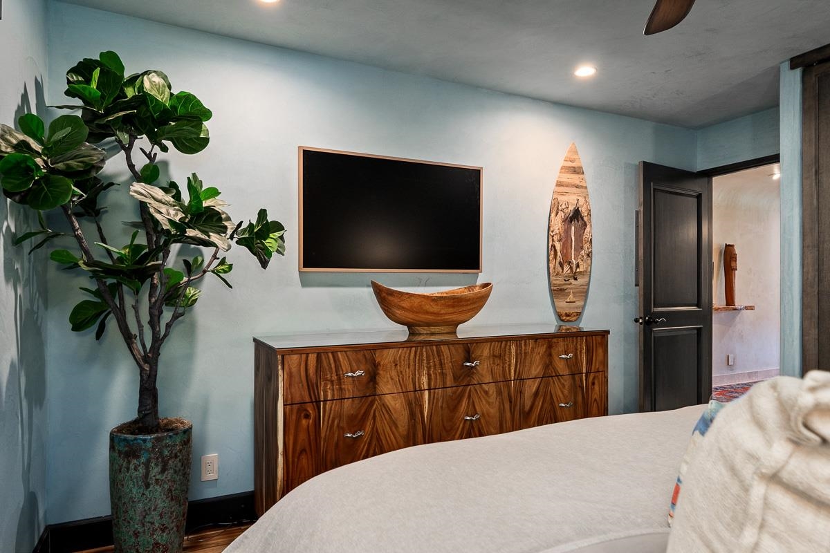 Bedroom with a wood dresser, wall-mounted television, potted tree, and surfboard-shaped wall art.