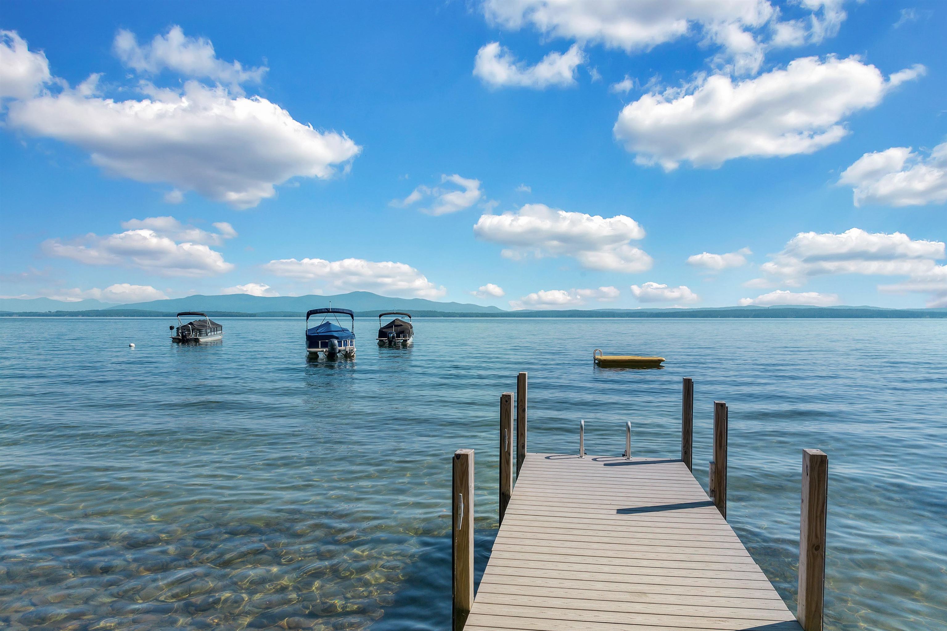 Wooden dock extends into a lake toward moored boats, with distant mountains across the water.