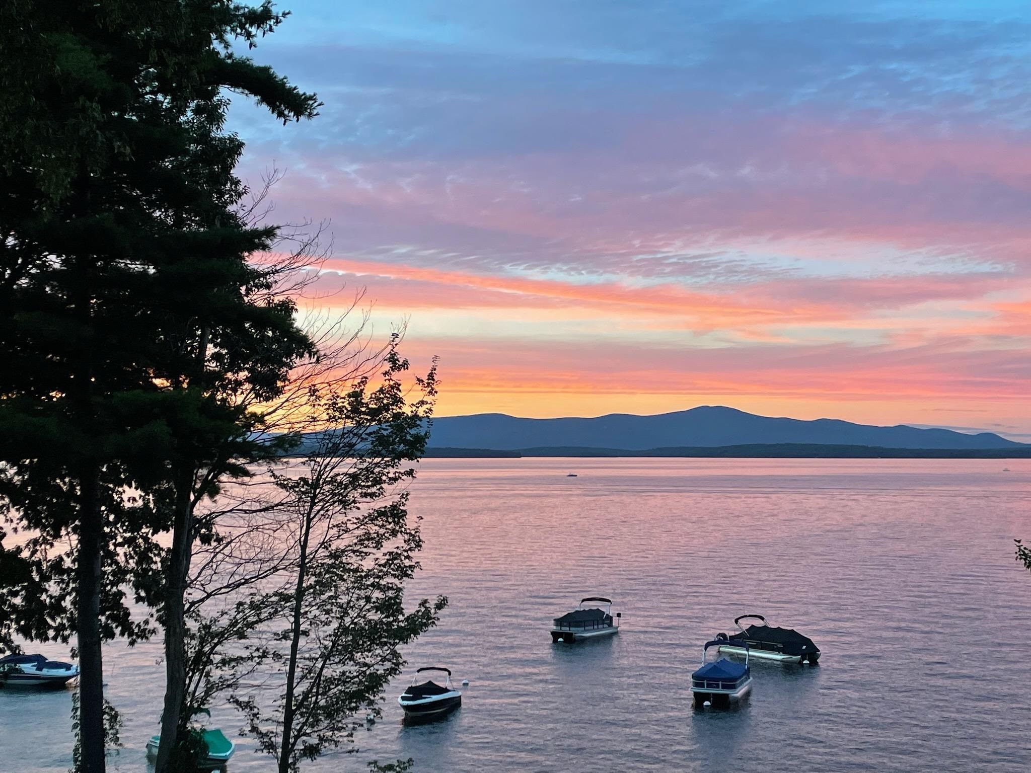 Lake at sunset with boats, silhouetted trees, and mountains across the water.