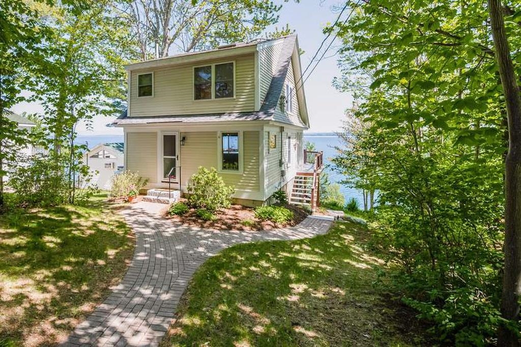 Two-story lakeside house with a curved brick walkway and leafy trees.