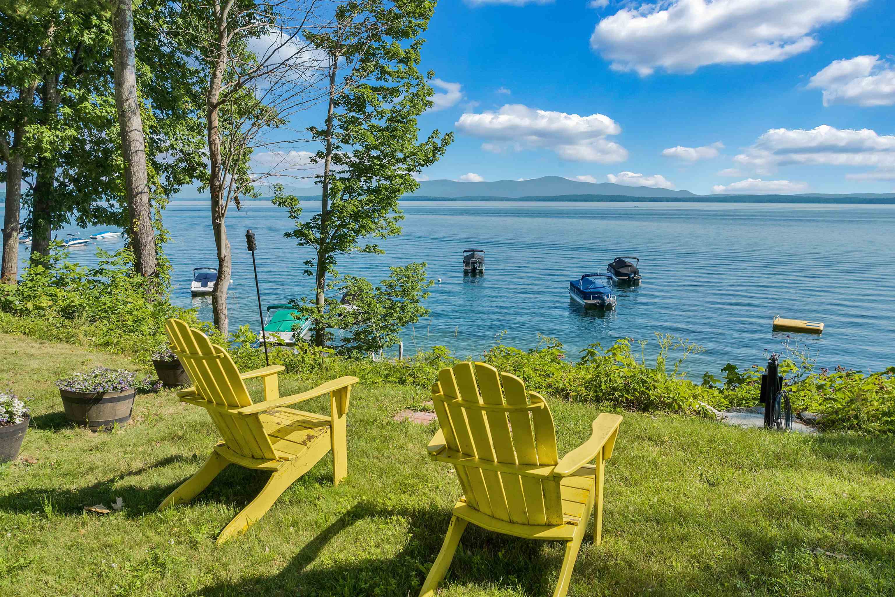 Two yellow Adirondack chairs overlook a lake dotted with boats, with mountains on the horizon.