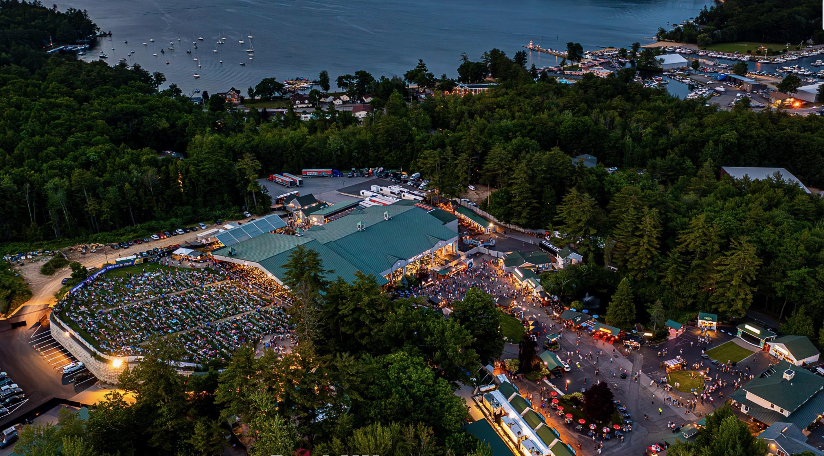 Aerial view of a crowded outdoor venue beside a lake, with green-roofed buildings and boats in the harbor.