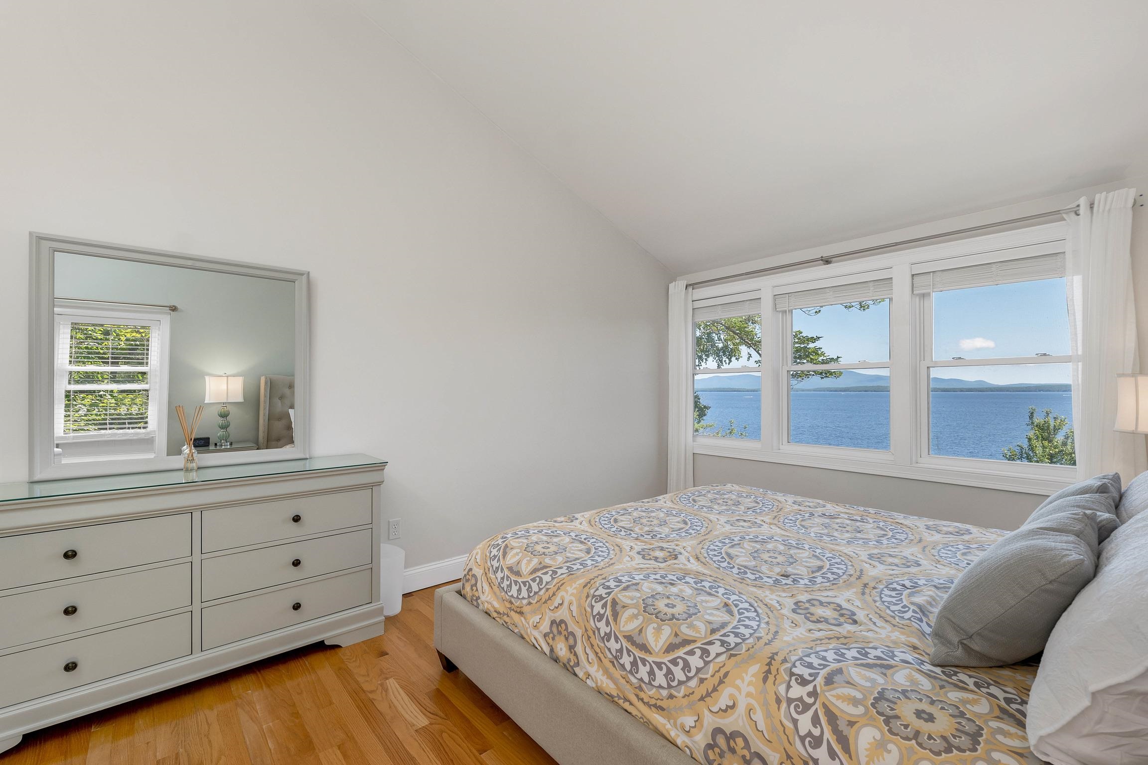 Bedroom with a patterned bedspread and lake views through three large windows.