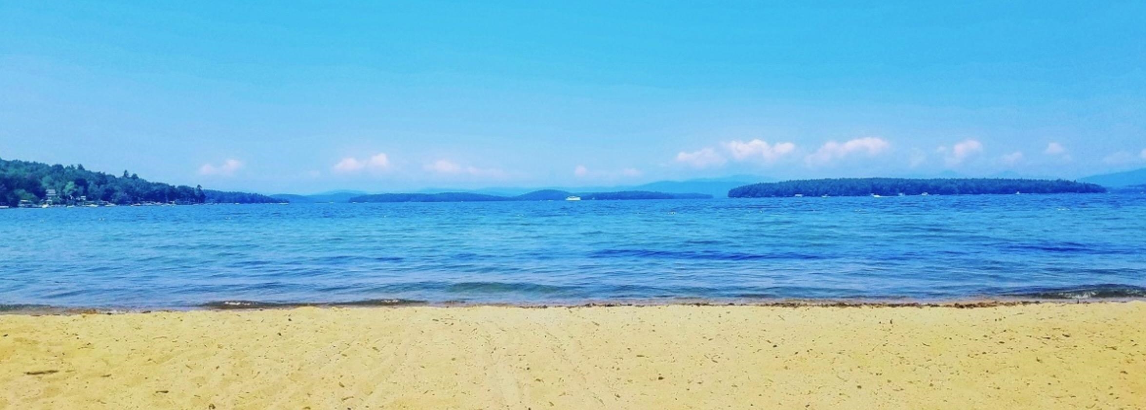 Sandy beach and blue lake with wooded shores and distant hills.