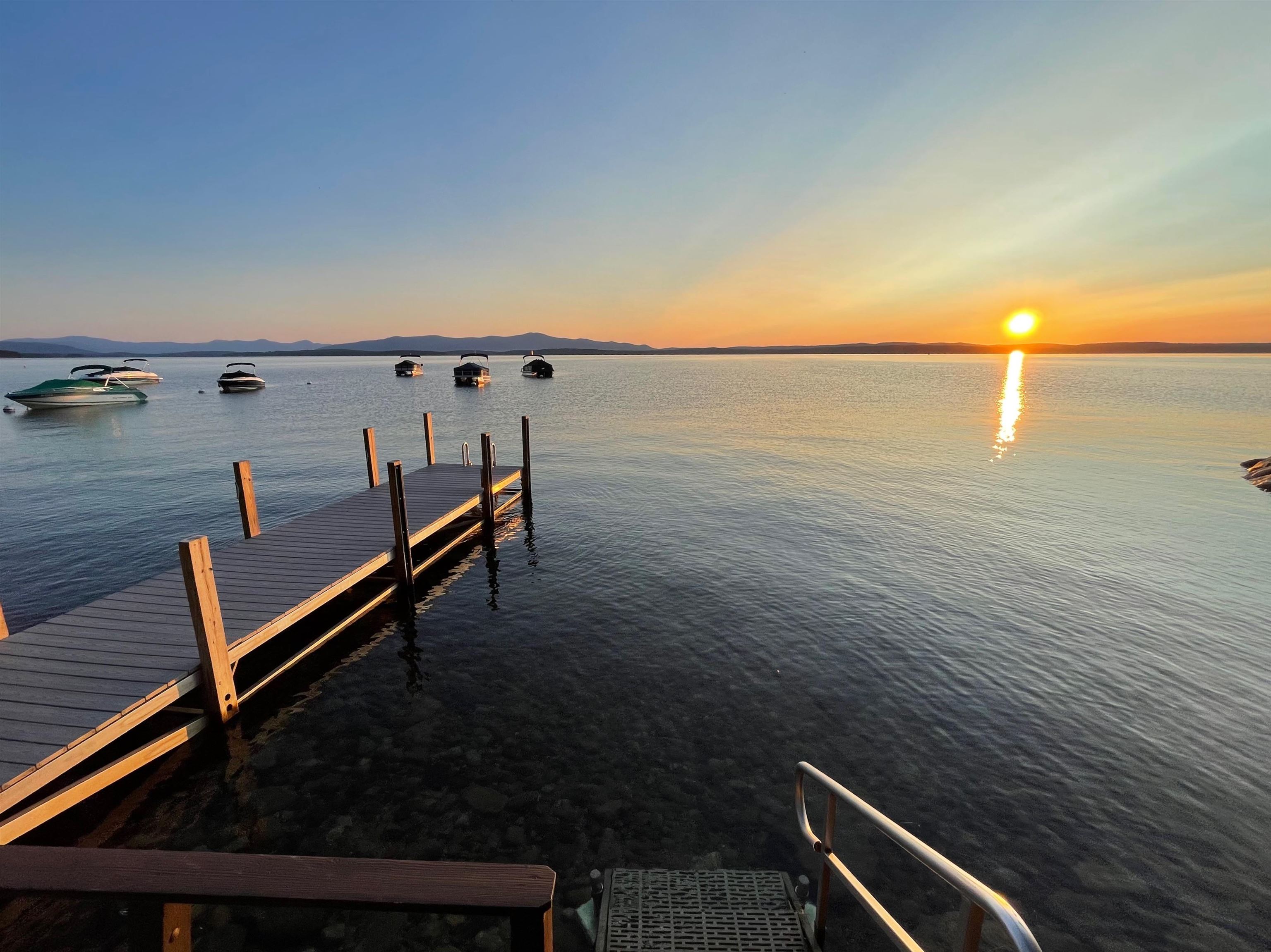 Wooden dock on a calm lake at sunset, with moored boats and distant mountains.