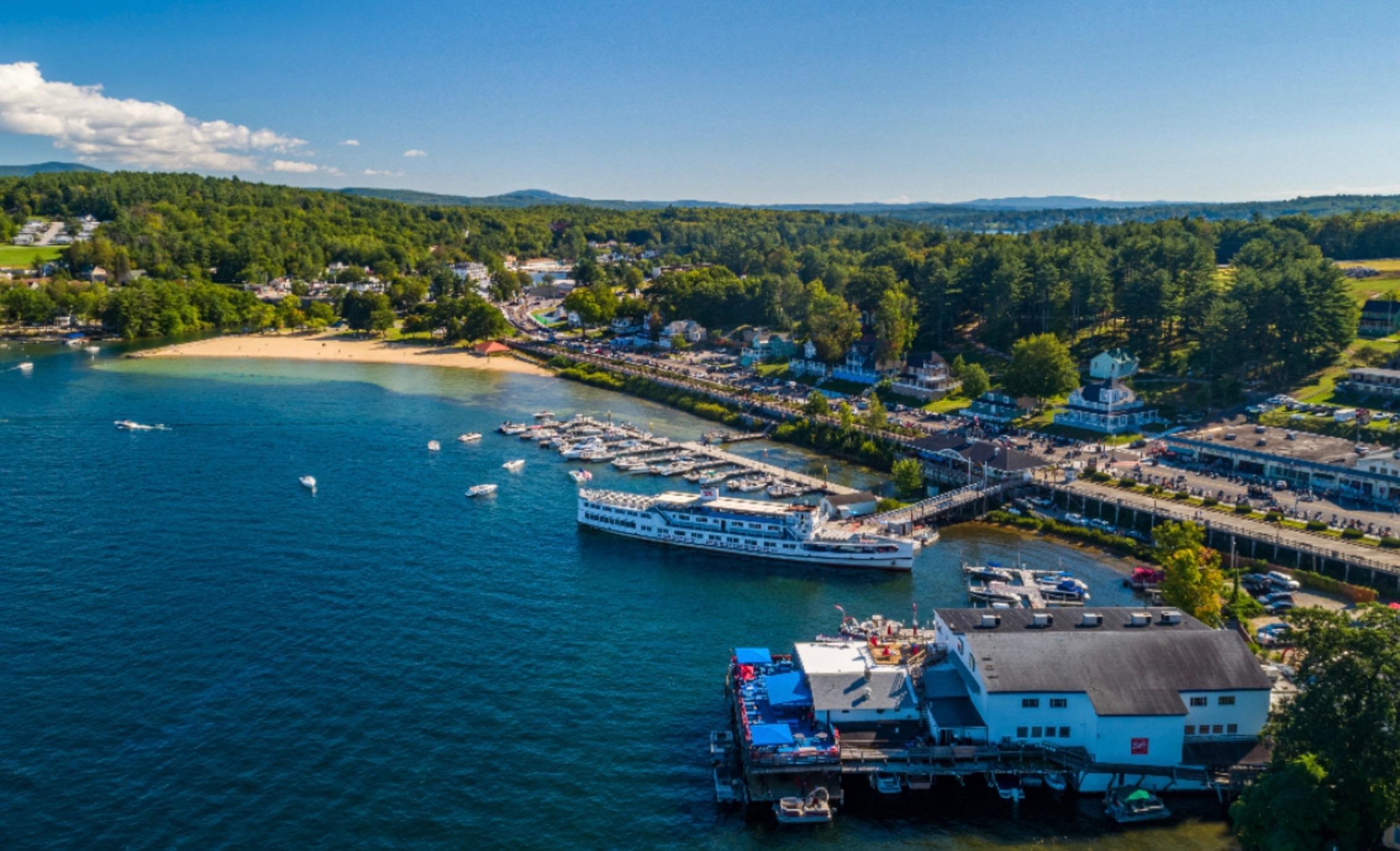 Aerial view of a lakeside marina, sandy beach, boats, and wooded shoreline.