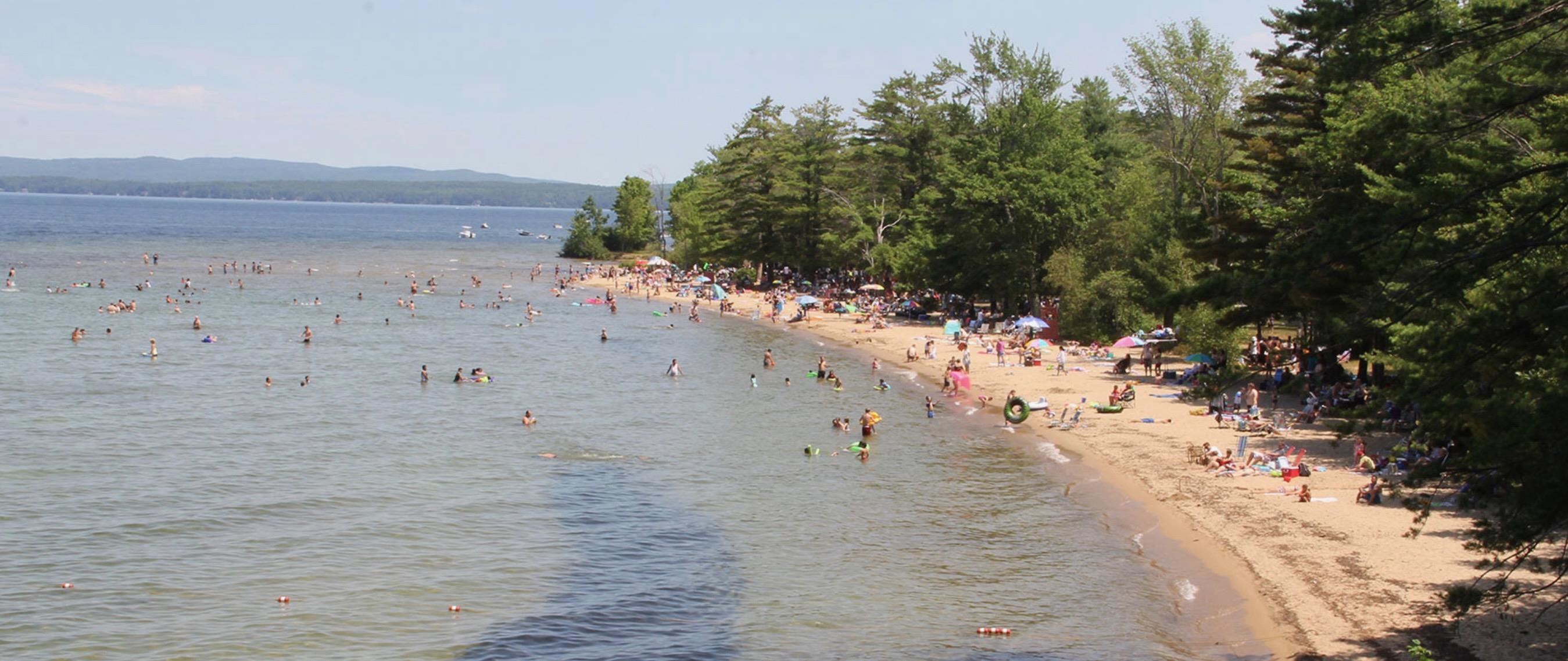 Crowded lakeside beach with swimmers, a sandy tree-lined shore, and forested hills across the water.