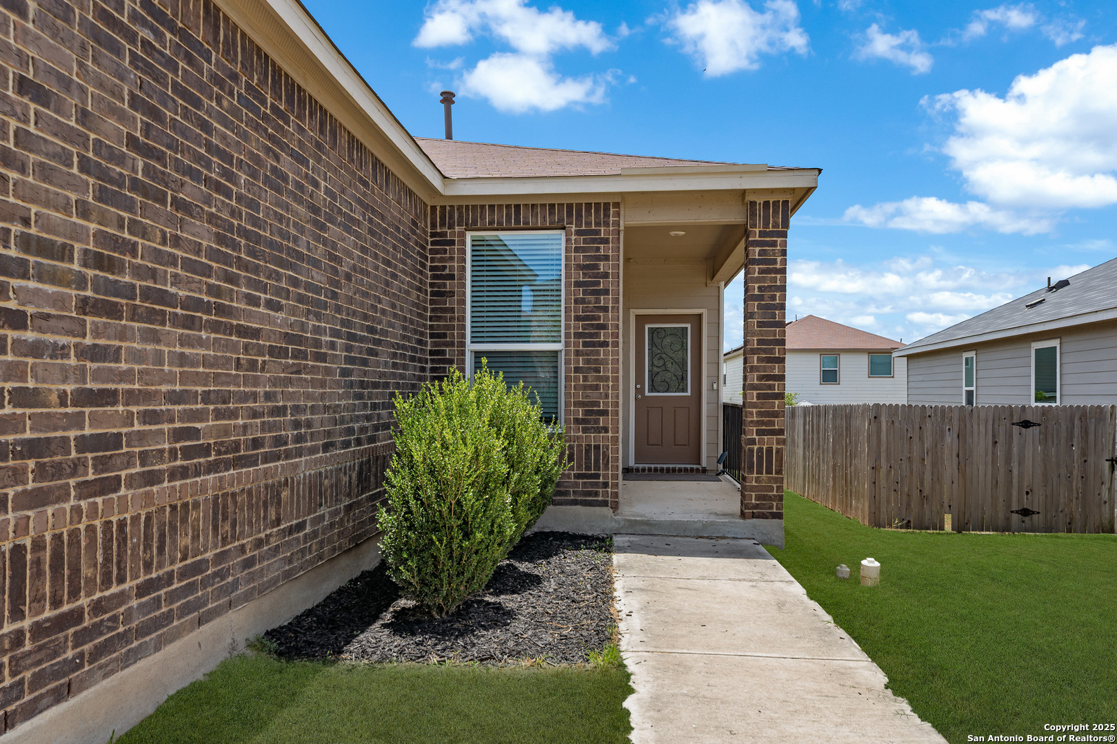 Brick home entrance with a covered porch, concrete walkway, shrub bed, and fenced lawn.