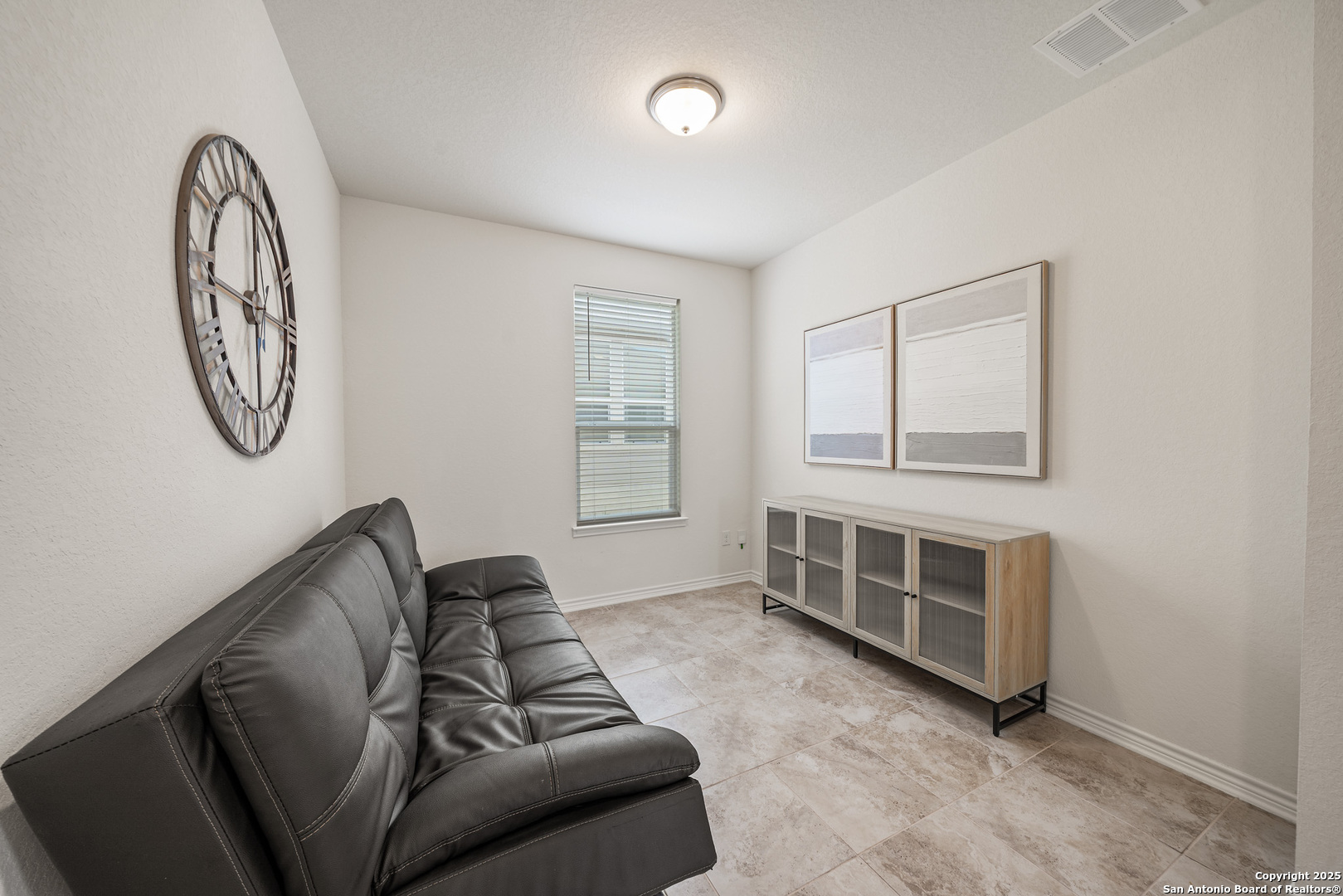 Living room with a dark leather sofa, tiled floor, large wall clock, and glass-front console.