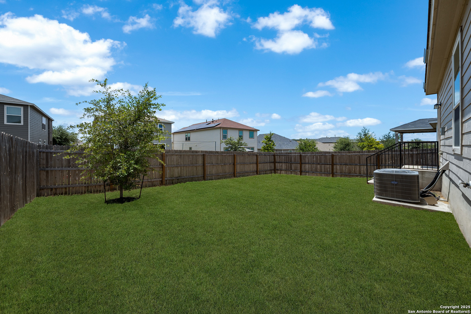 Fenced backyard with a broad lawn, young tree, and raised patio beside the house.