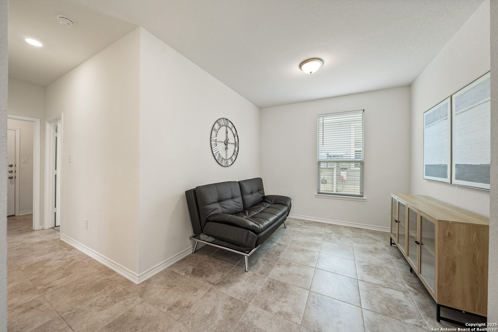 Living room with a black leather loveseat, beige tile floors, and a large wall clock.