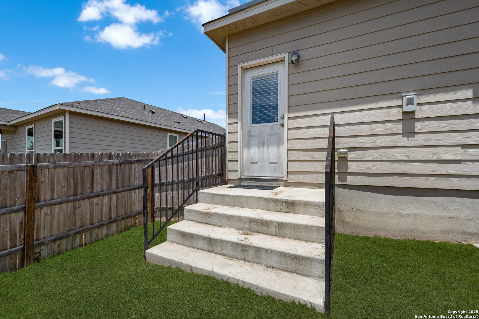 Home entrance with concrete steps and black railings beside a fenced grassy yard.