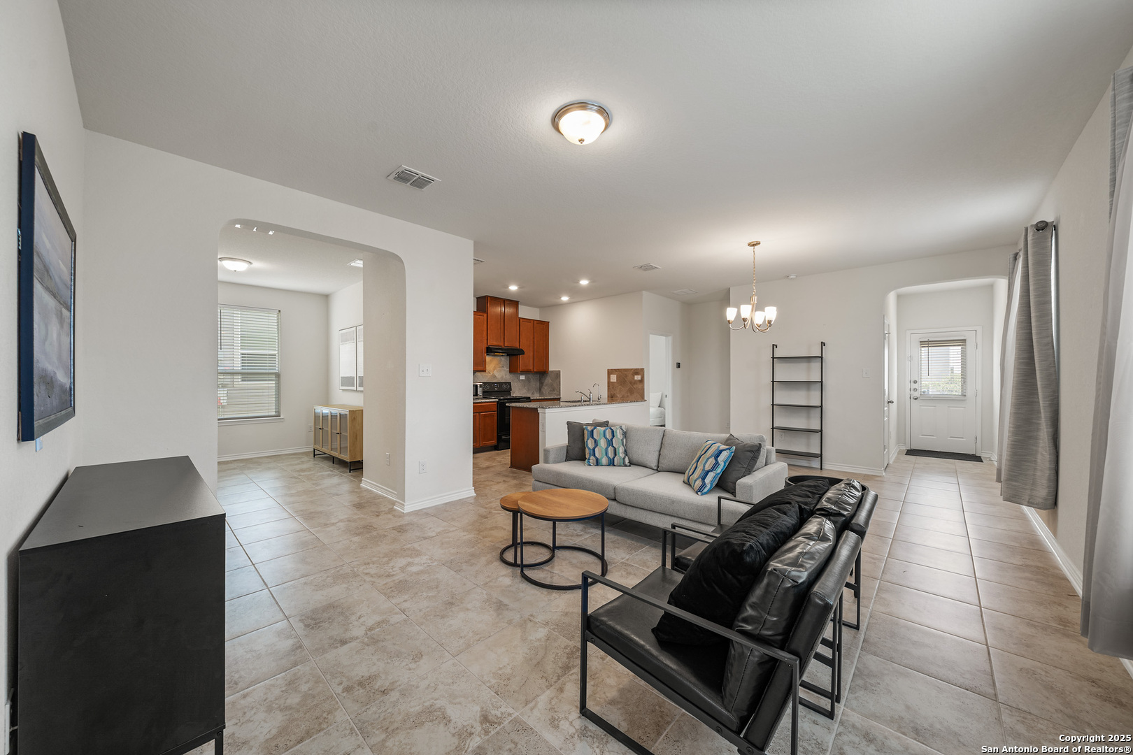 Open-plan living room with gray sectional, black chairs, tiled floors, and kitchen in view.