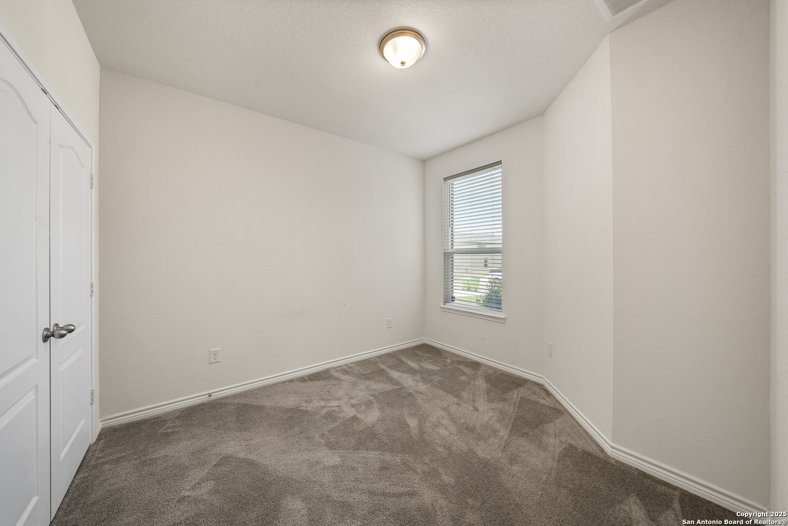 Empty bedroom with beige carpet, a window with blinds, and a ceiling light.