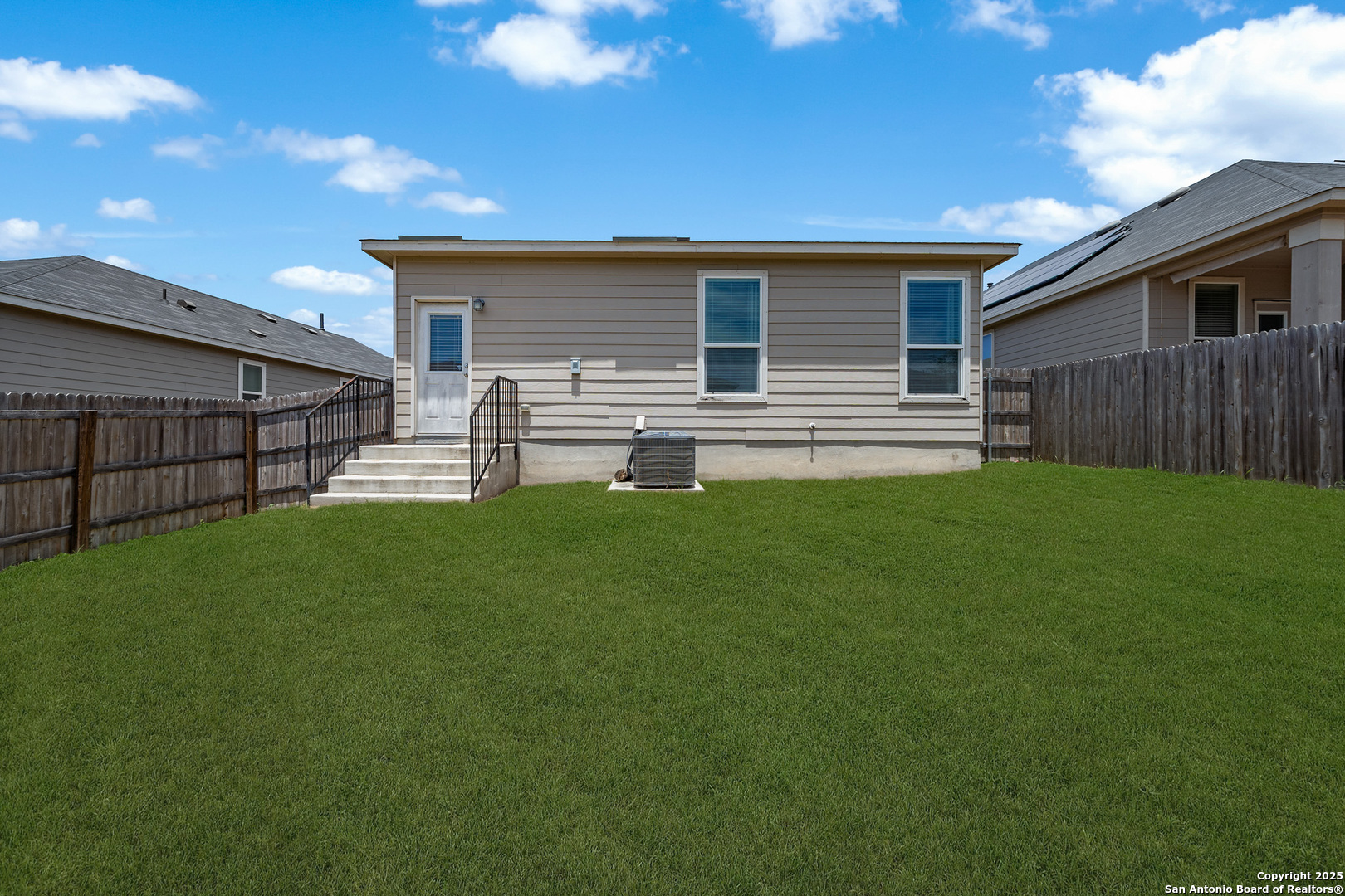 Back of a beige house with fenced green lawn, concrete steps, black railings, and an outdoor air-conditioning unit.