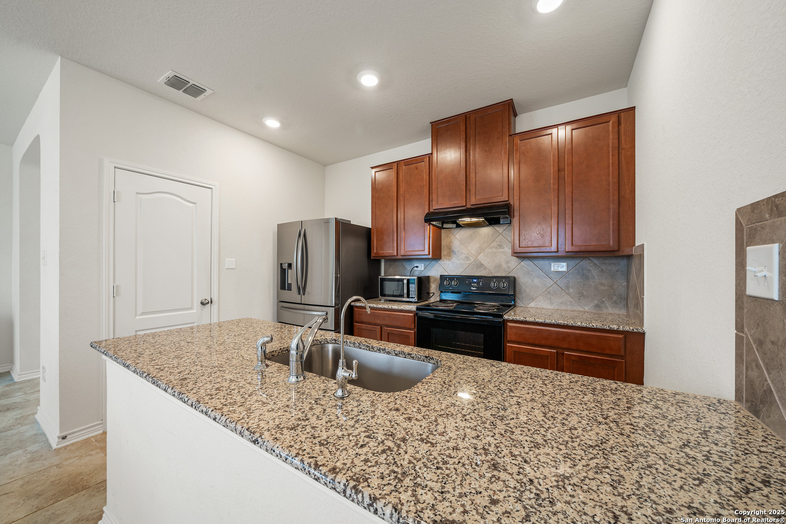 Kitchen with reddish-brown cabinets, granite breakfast bar, stainless refrigerator, and tiled backsplash.