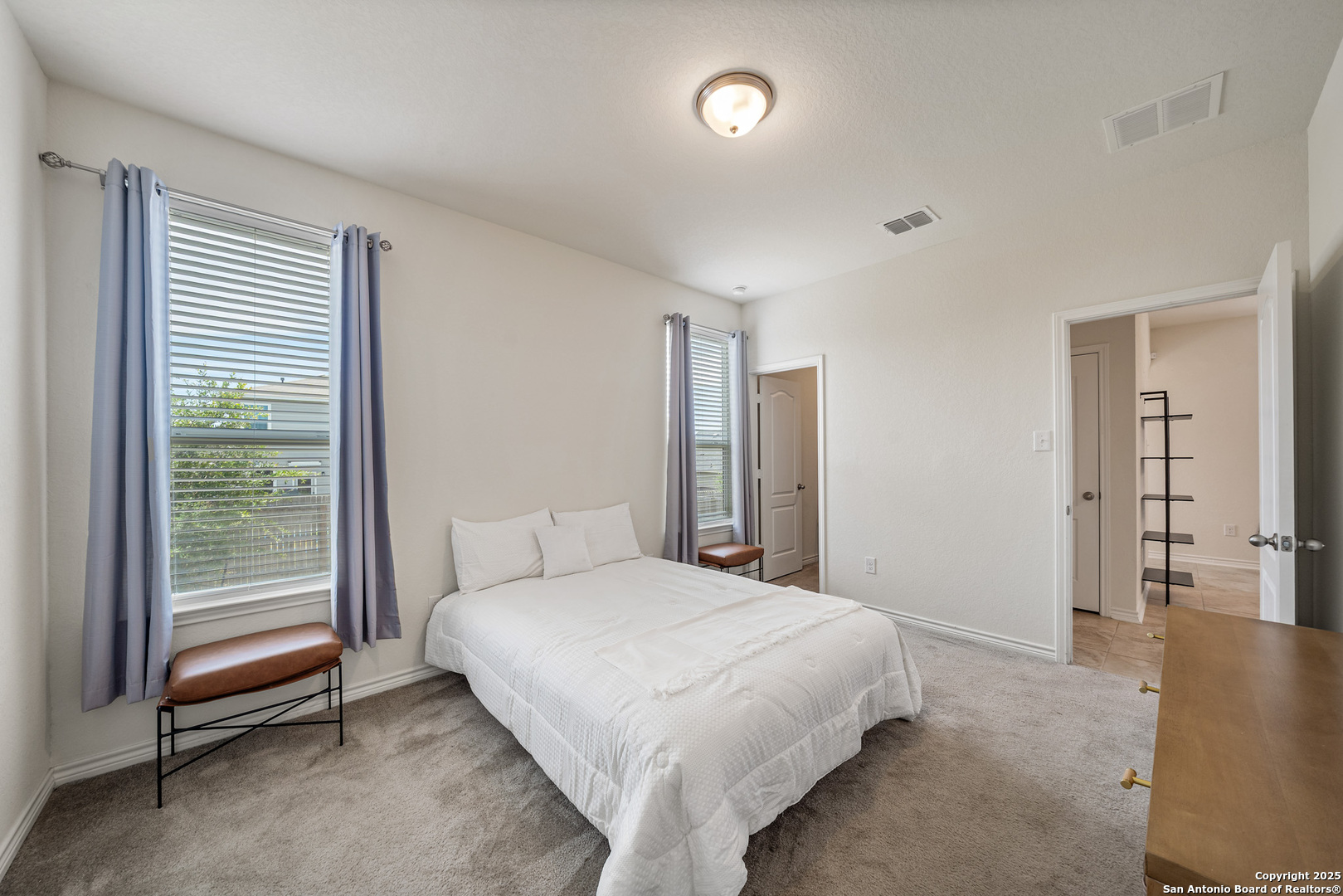 Bedroom with a white bed, gray carpet, two curtained windows, and a brown bench.