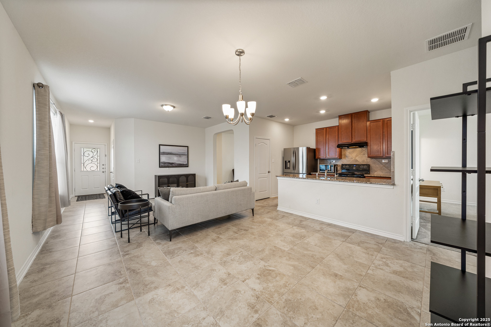 Open-plan living room and kitchen with beige tile floors, a gray sofa, and reddish-brown kitchen cabinets.