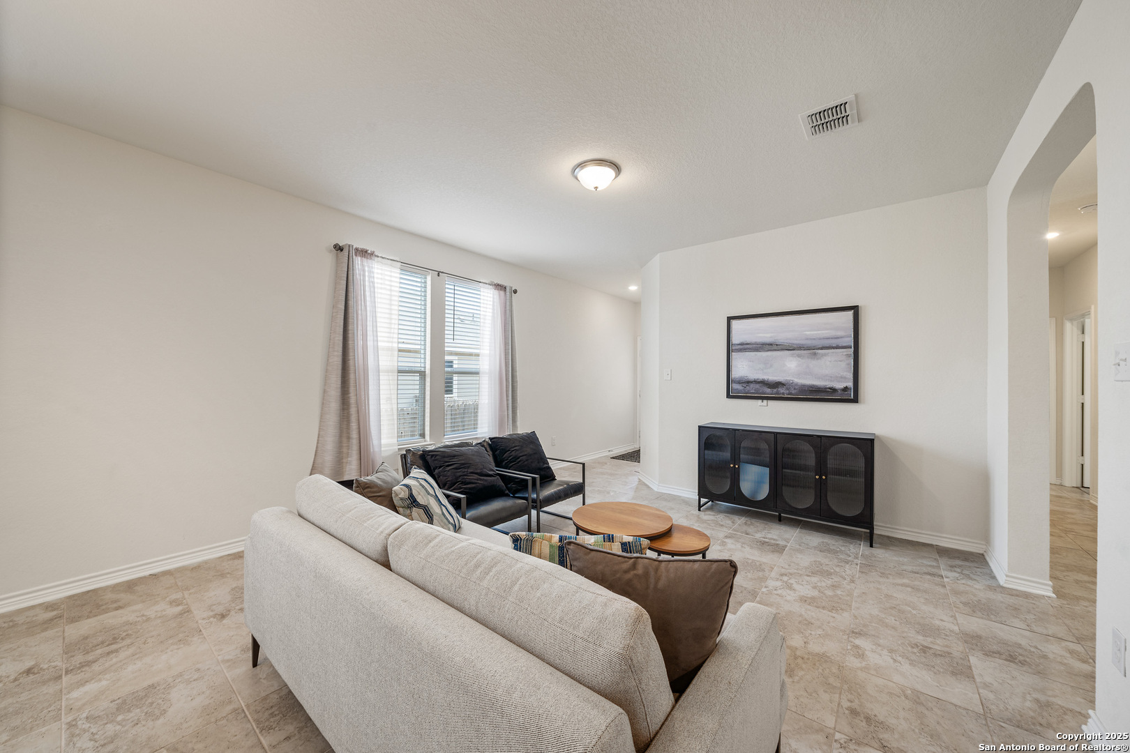 Staged living room with tile floors, a light sofa, black loveseat, and arched hallway opening.