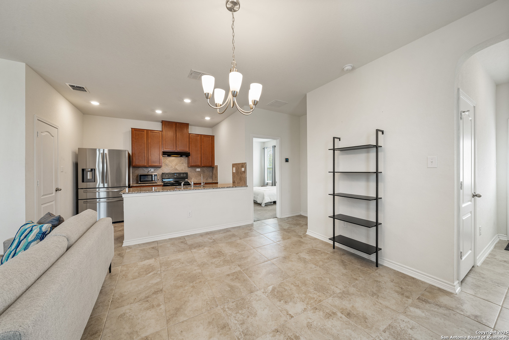 Open living room and kitchen with tiled floors, wood cabinets, stainless steel appliances, and black shelving.