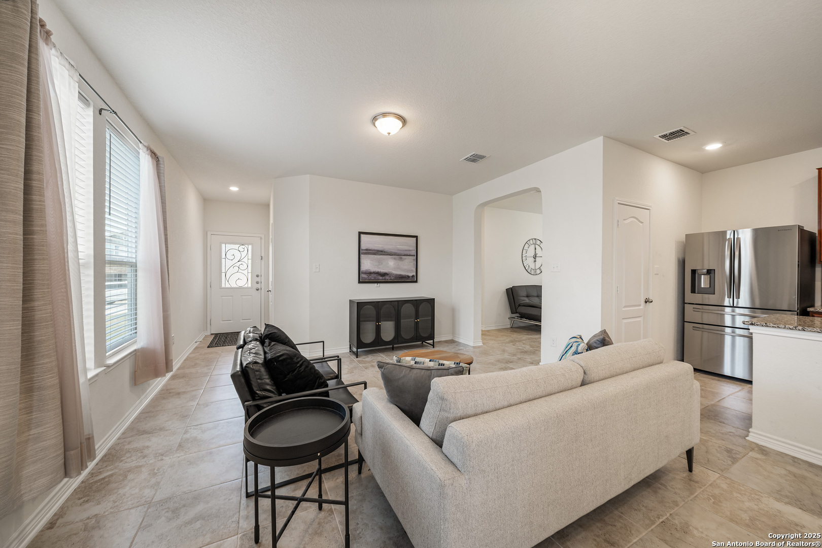 Furnished living room with large curtained windows, beige tile floors, and an open kitchen.