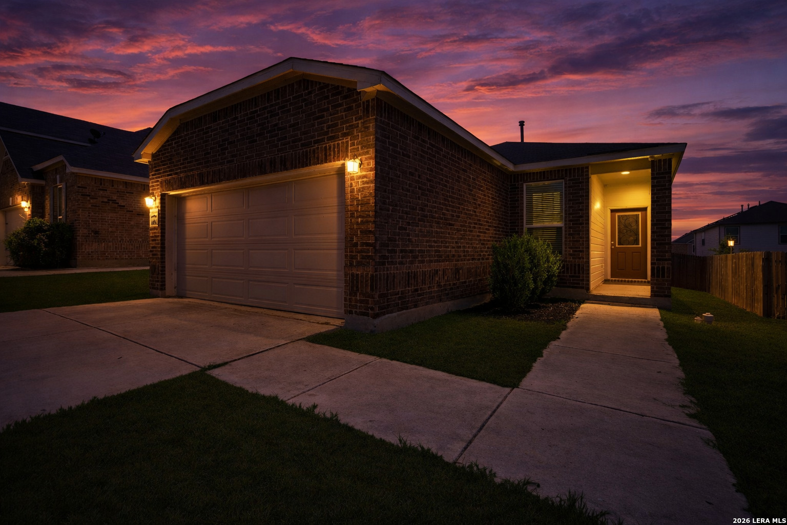 Brick house at dusk with a wide garage, illuminated entry, and concrete driveway beneath a colorful sunset sky.