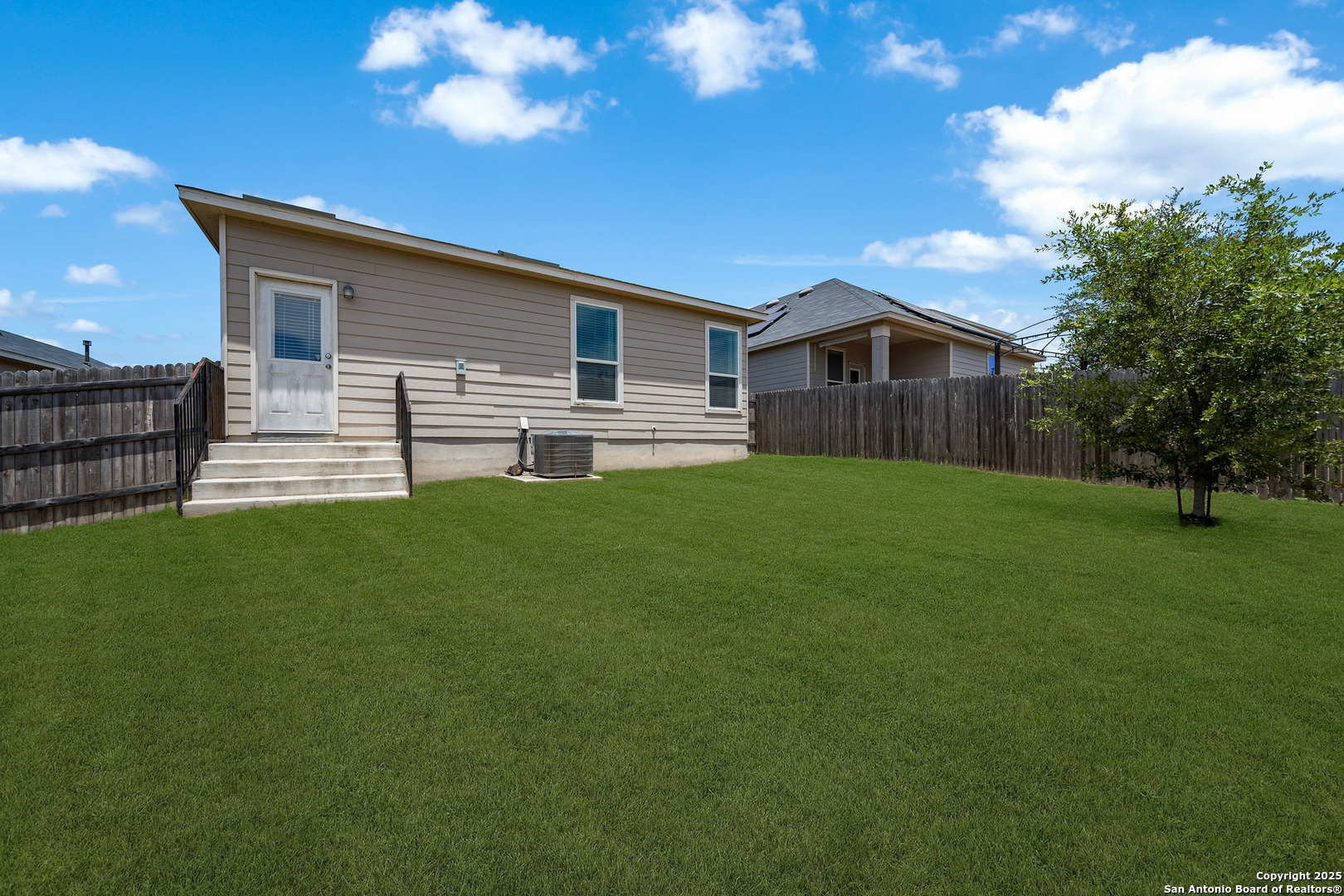 Back of a beige-sided house with white steps, a fenced lawn, and a small tree.
