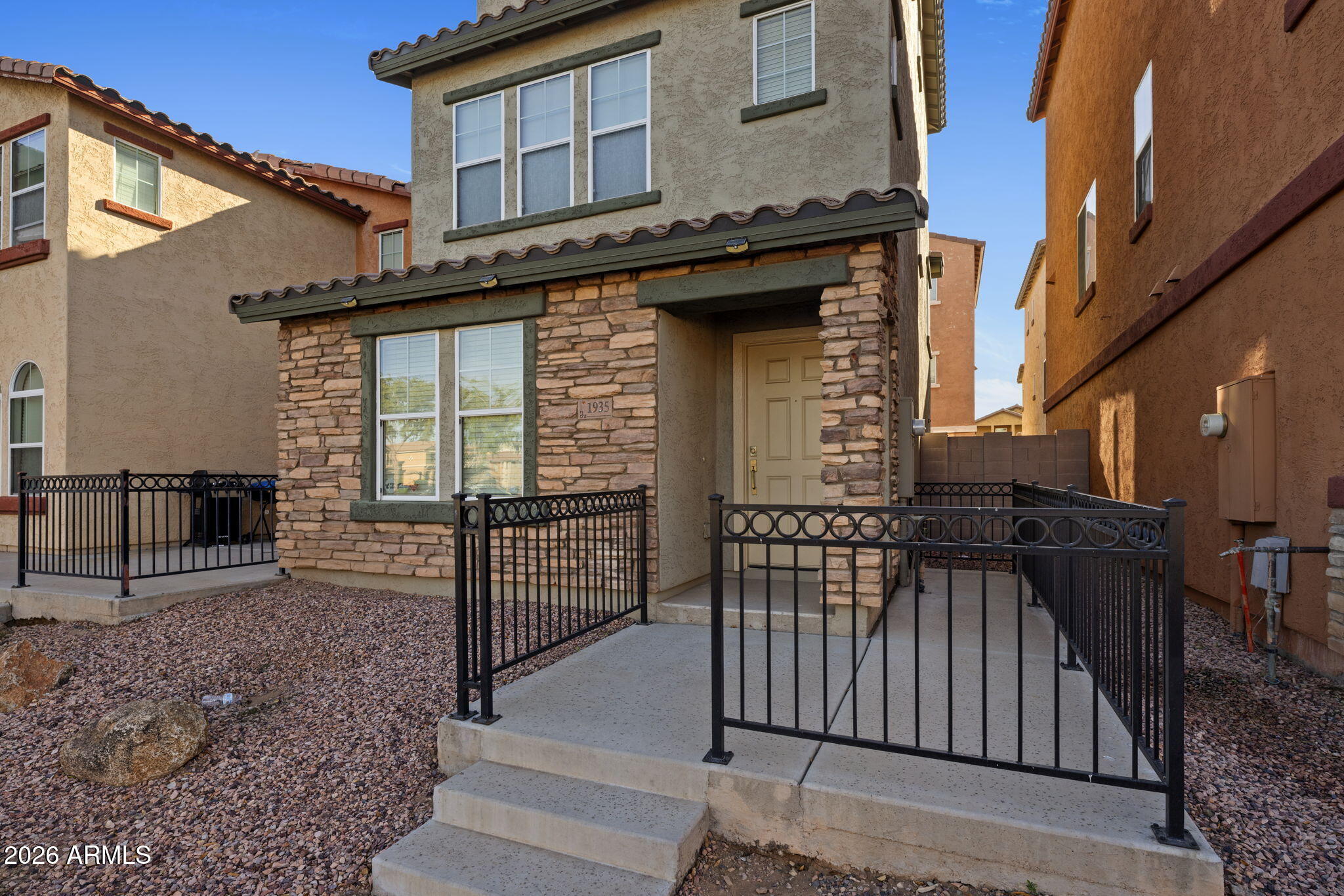 Two-story stucco home with stone veneer, black metal entry railings, and a raised concrete walkway.