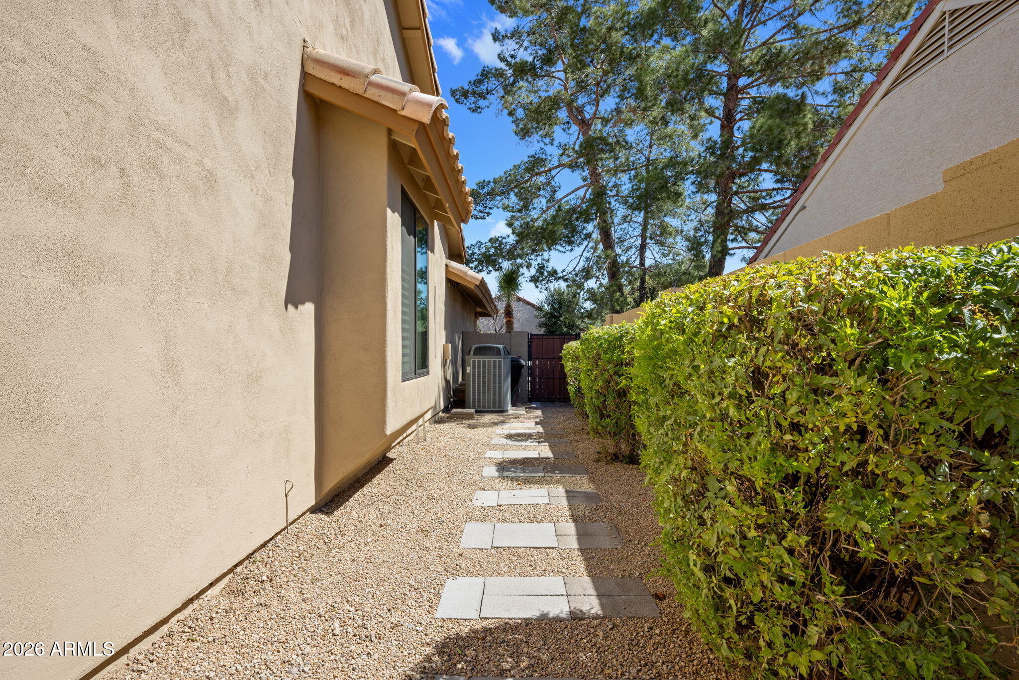 Home side yard with a gravel path, stepping stones, tall hedge, and outdoor air-conditioning unit.