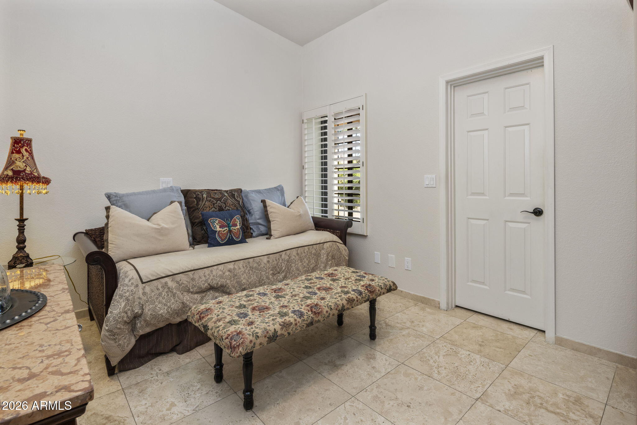 Furnished sitting area with a daybed-style sofa, floral bench, shuttered window, and beige tile floor.