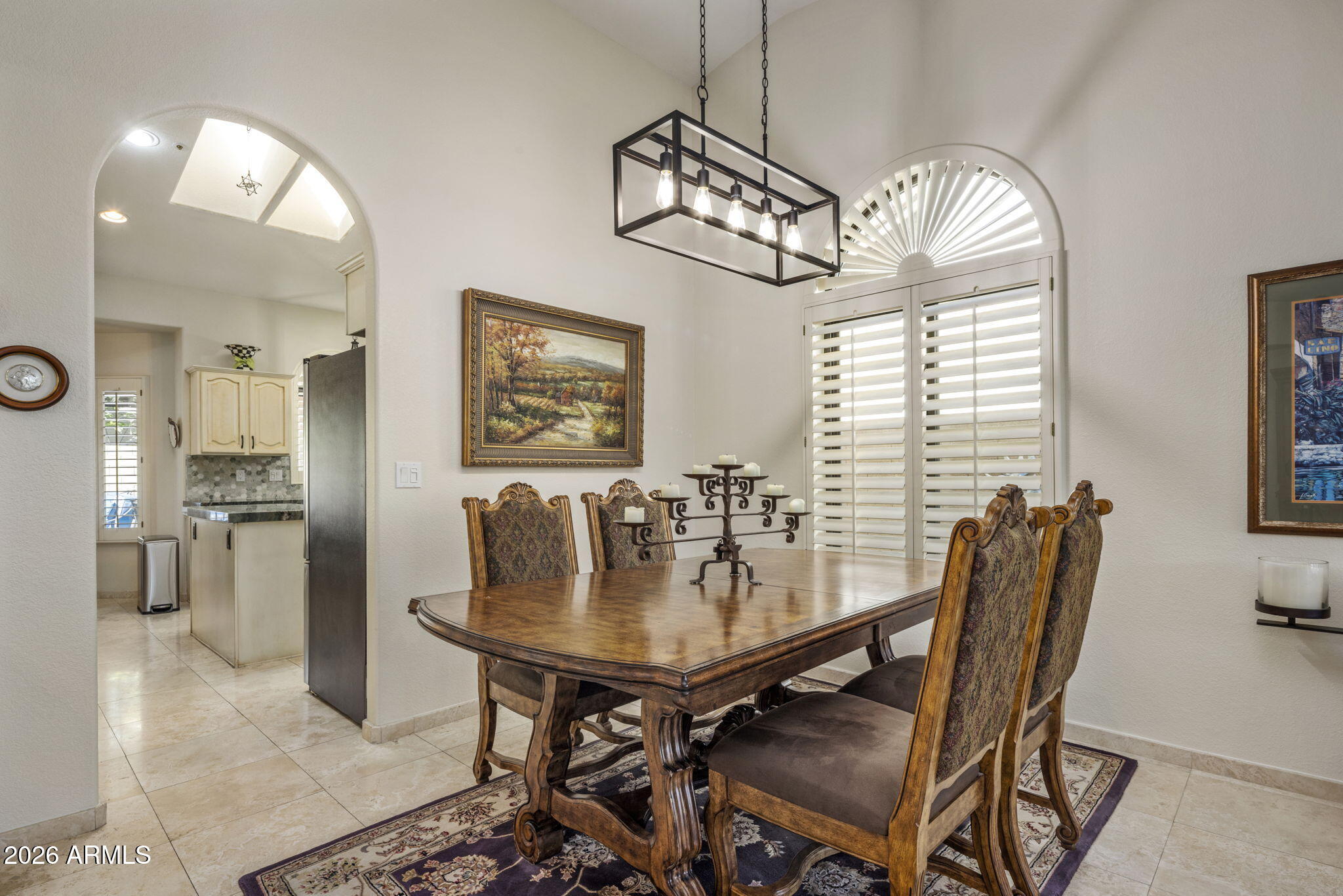 Furnished dining room with ornate wood table, arched plantation-shutter window, and kitchen opening.