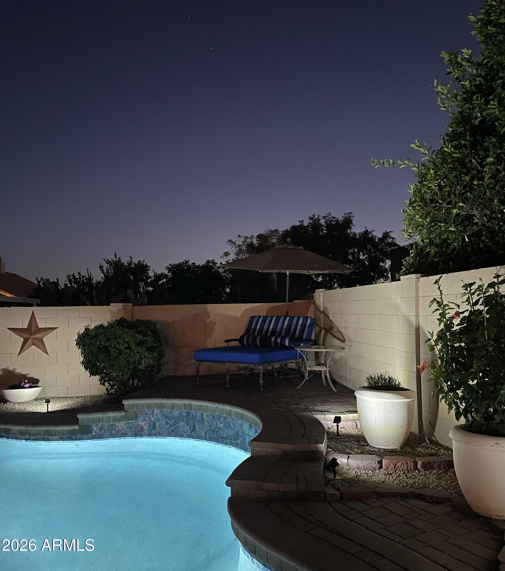 Blue-lit backyard pool beside a paved patio with an umbrella-shaded striped lounge chair at night.