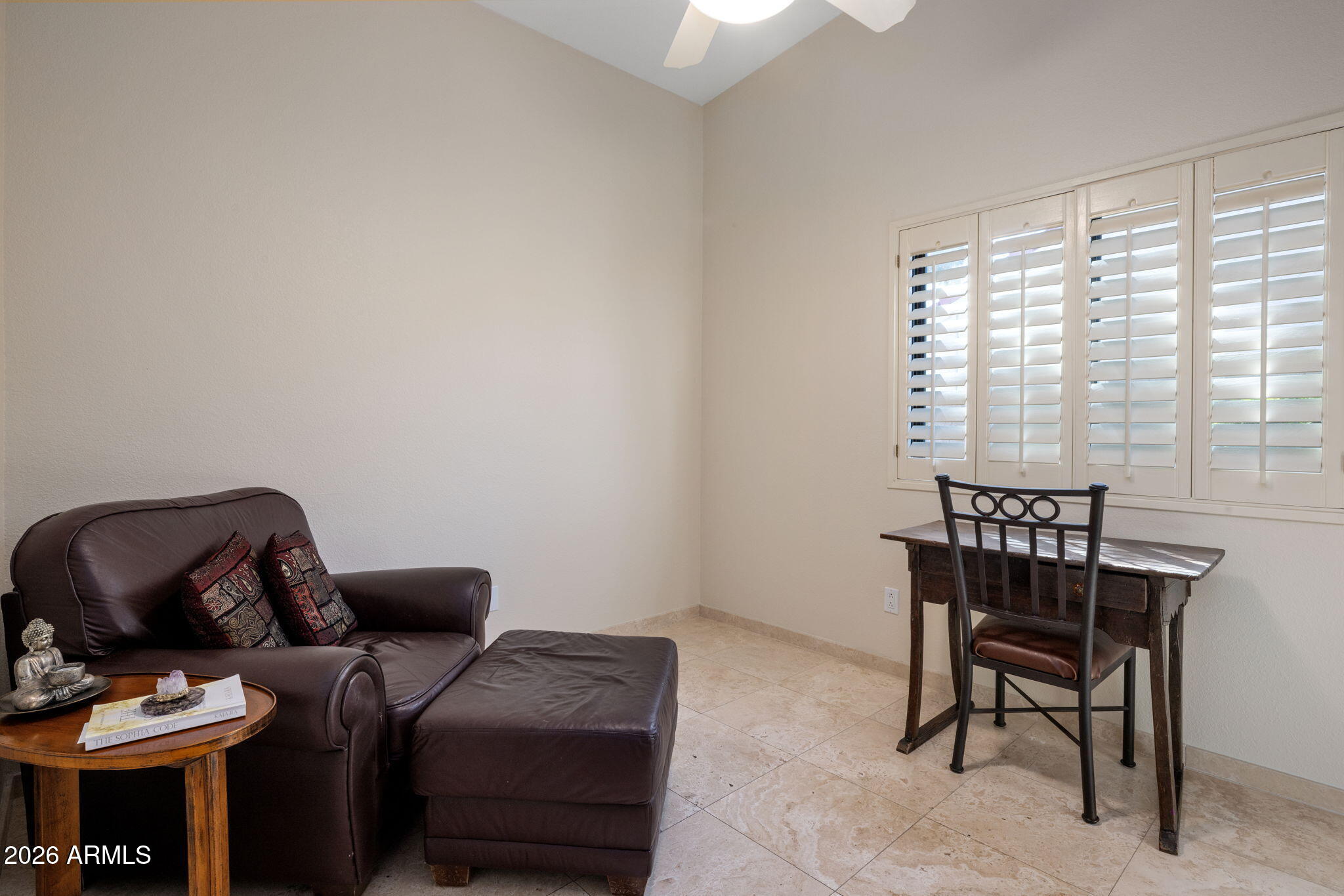 Furnished sitting room with a leather loveseat, ottoman, writing desk, and plantation-shuttered window.